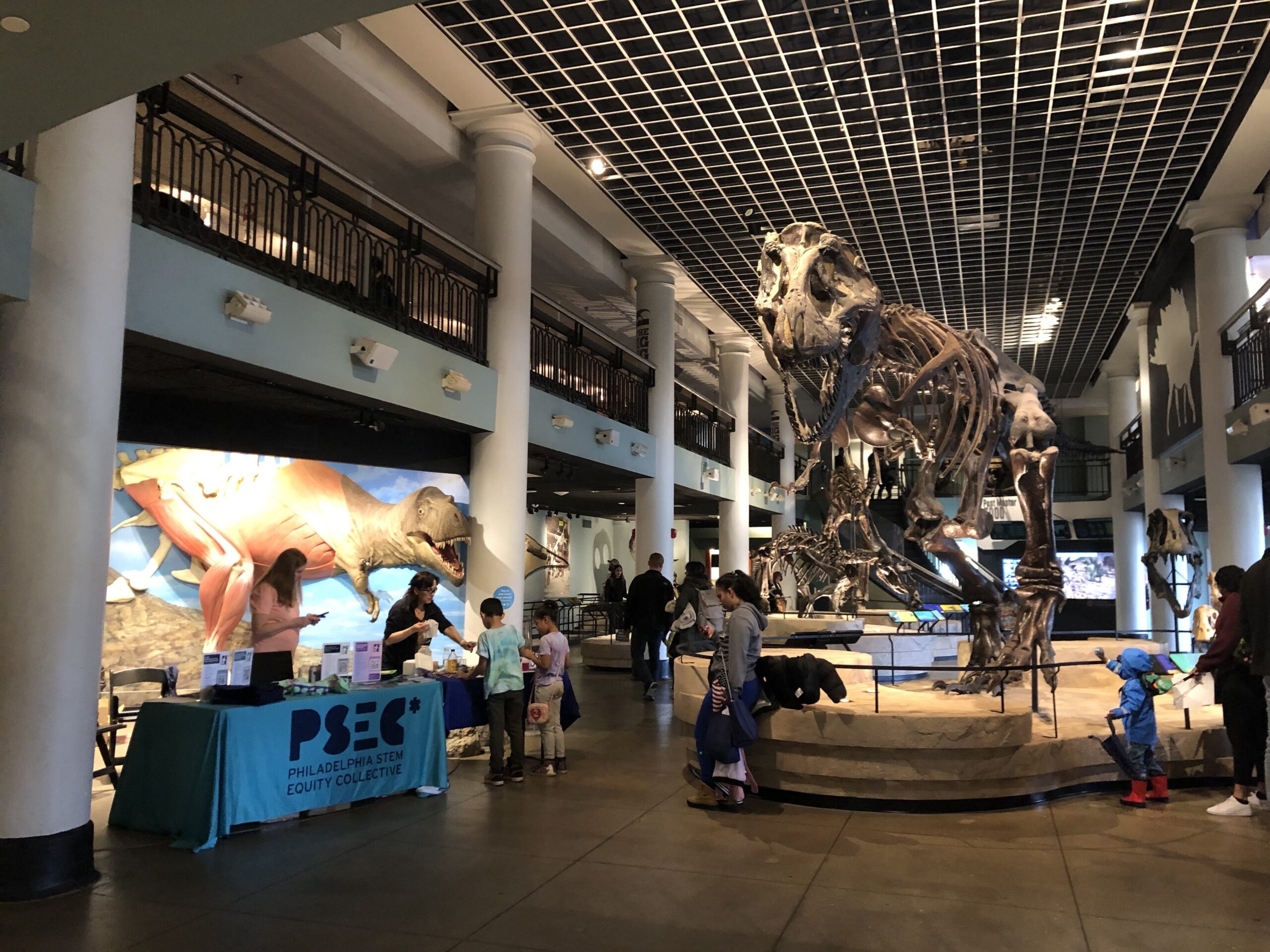 Museum lobby with a large dinosaur skeleton on display. People interact at a table and view exhibits. Art of dinosaurs decorates the walls.