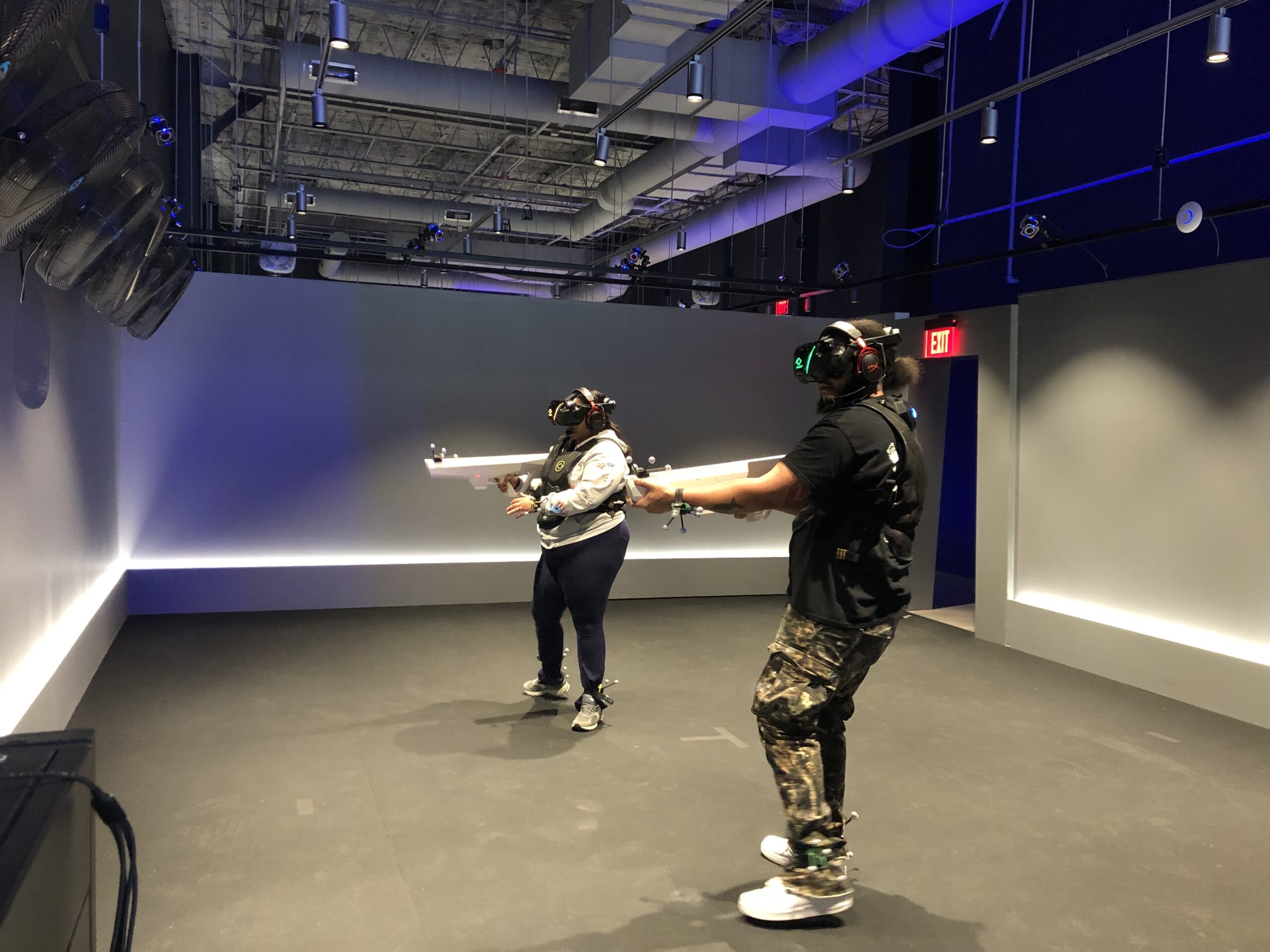 Two people wearing VR headsets and vests hold white controllers while standing in a dark, empty room with exposed ceilings and blue lighting.