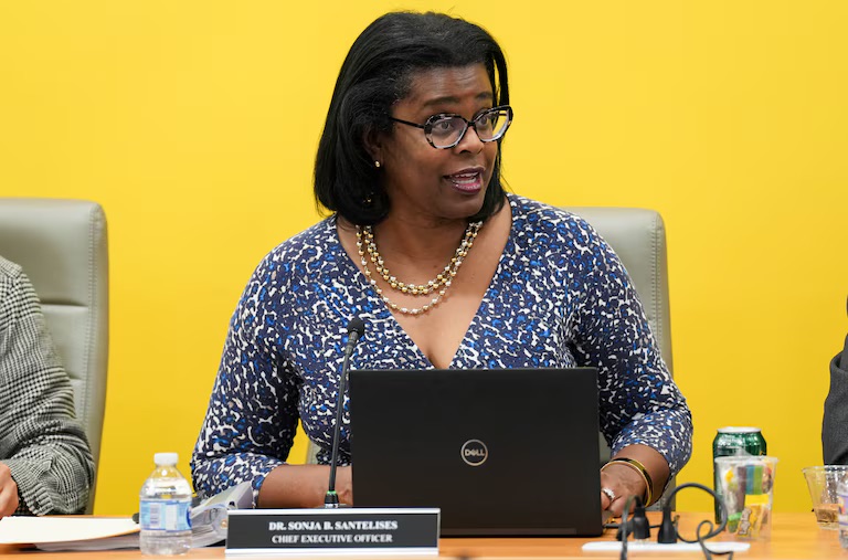 A woman sits at a conference table in front of a laptop, wearing glasses and a patterned blouse, with a nameplate reading "Dr. Sonja B. Santelises, Chief Executive Officer.