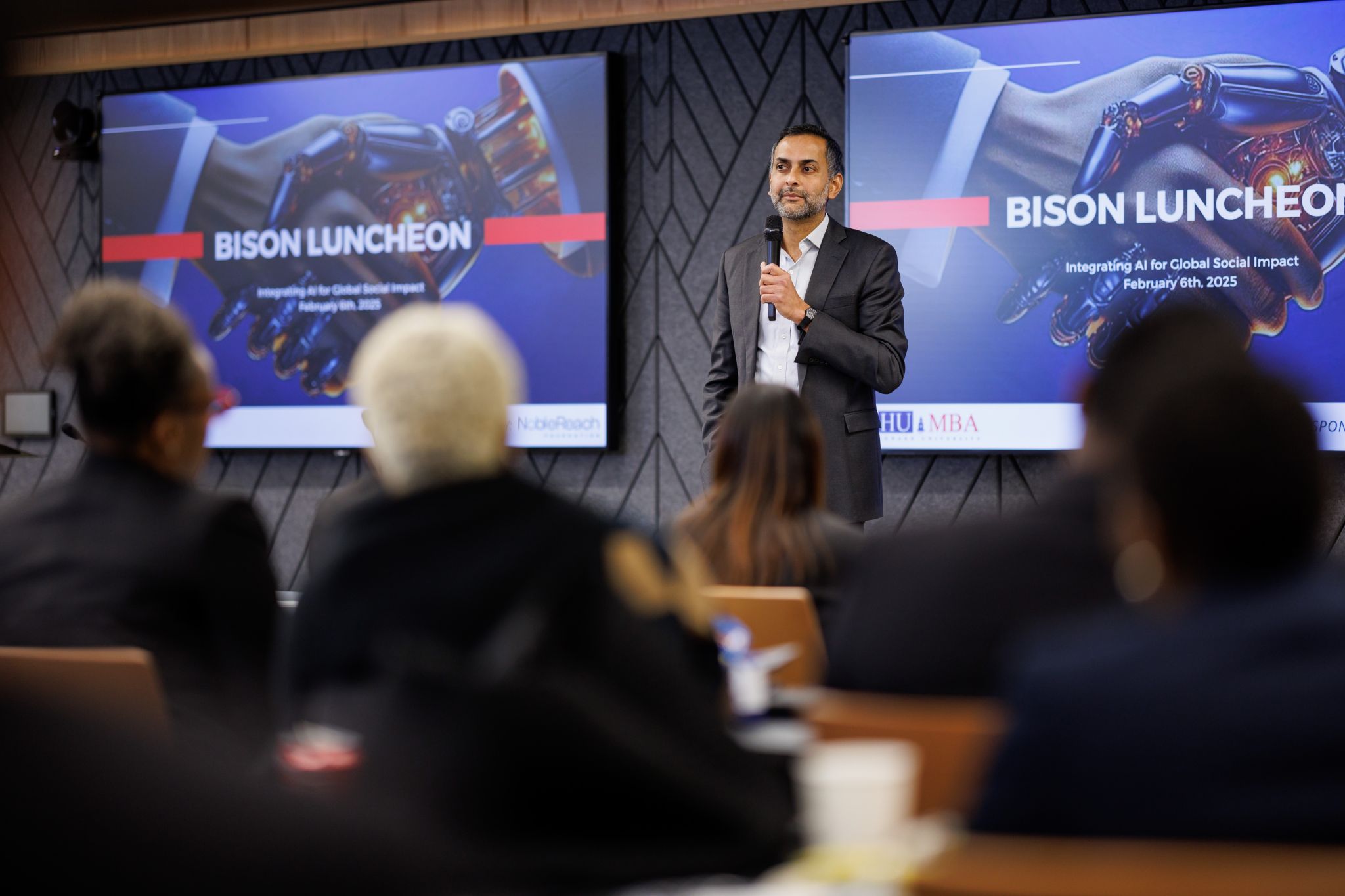 A man in a suit speaks into a microphone at a podium during a "Bison Luncheon" event, with attendees seated and a presentation displayed behind him.