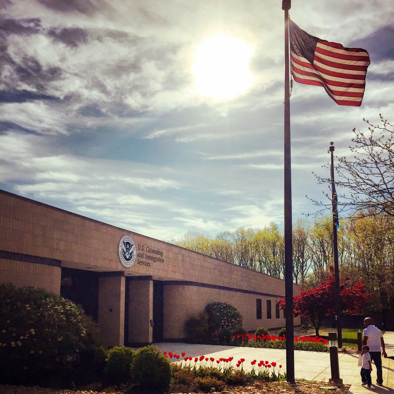 A U.S. Citizenship and Immigration Services building with an American flag in front, tulips blooming, and two people walking near the entrance on a sunny day.