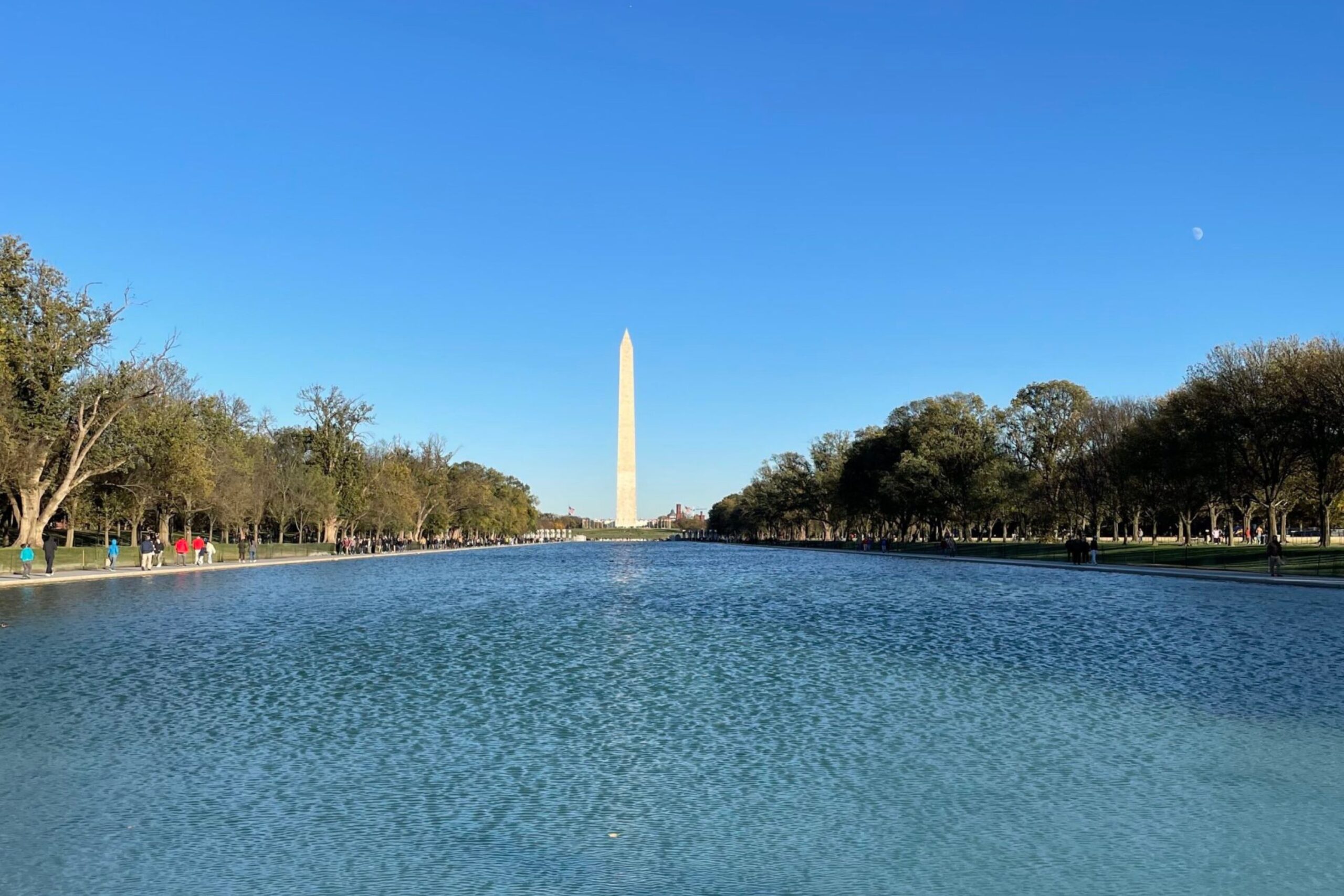 The Washington Monument stands in the distance, reflecting in the water of the National Mall’s reflecting pool under a clear blue sky.
