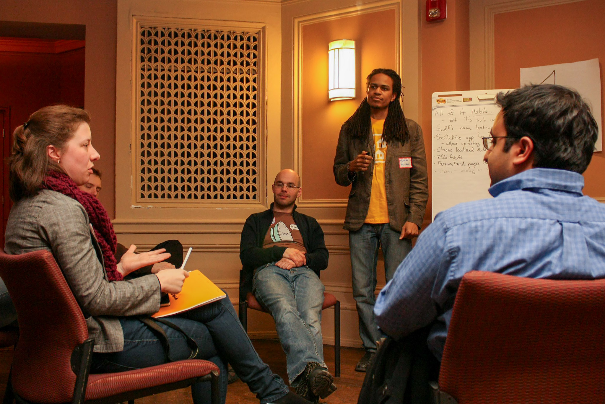 Five adults are engaged in a discussion in a small, indoor meeting room; one stands near a flip chart while others sit in chairs, listening attentively.