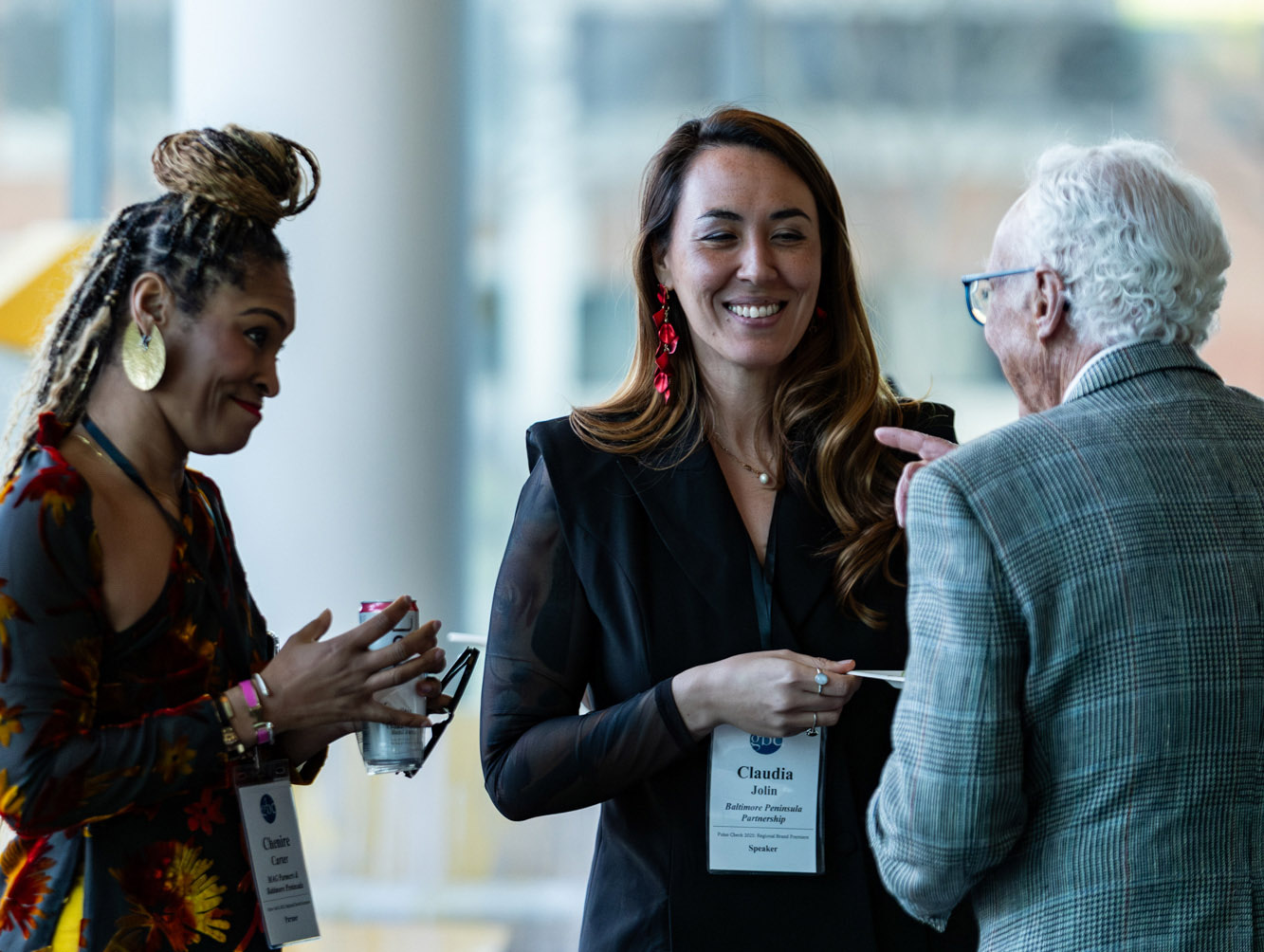 Three people converse indoors at an event. Two women with name badges face an older man in glasses and a checked jacket. One woman holds a drink; all appear engaged in conversation.