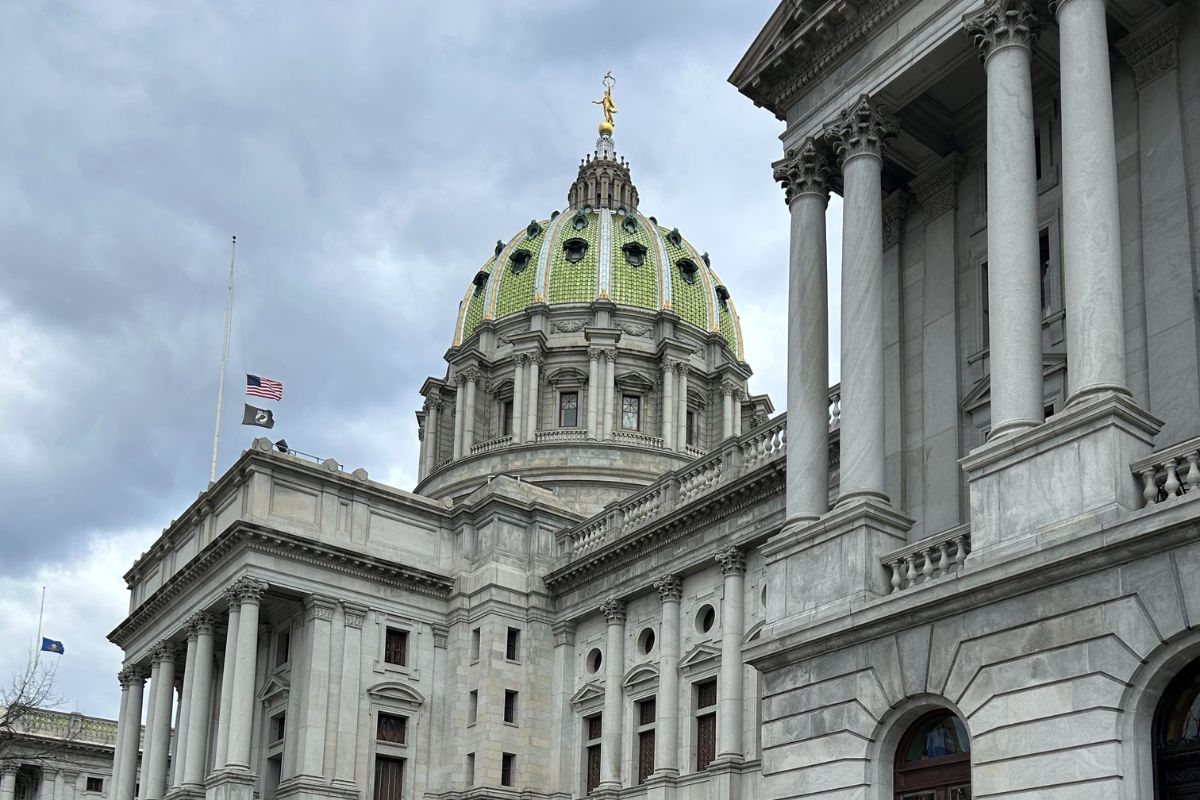 The Pennsylvania State Capitol building with a green dome, columns, and two flags flying at half-staff under a cloudy sky.