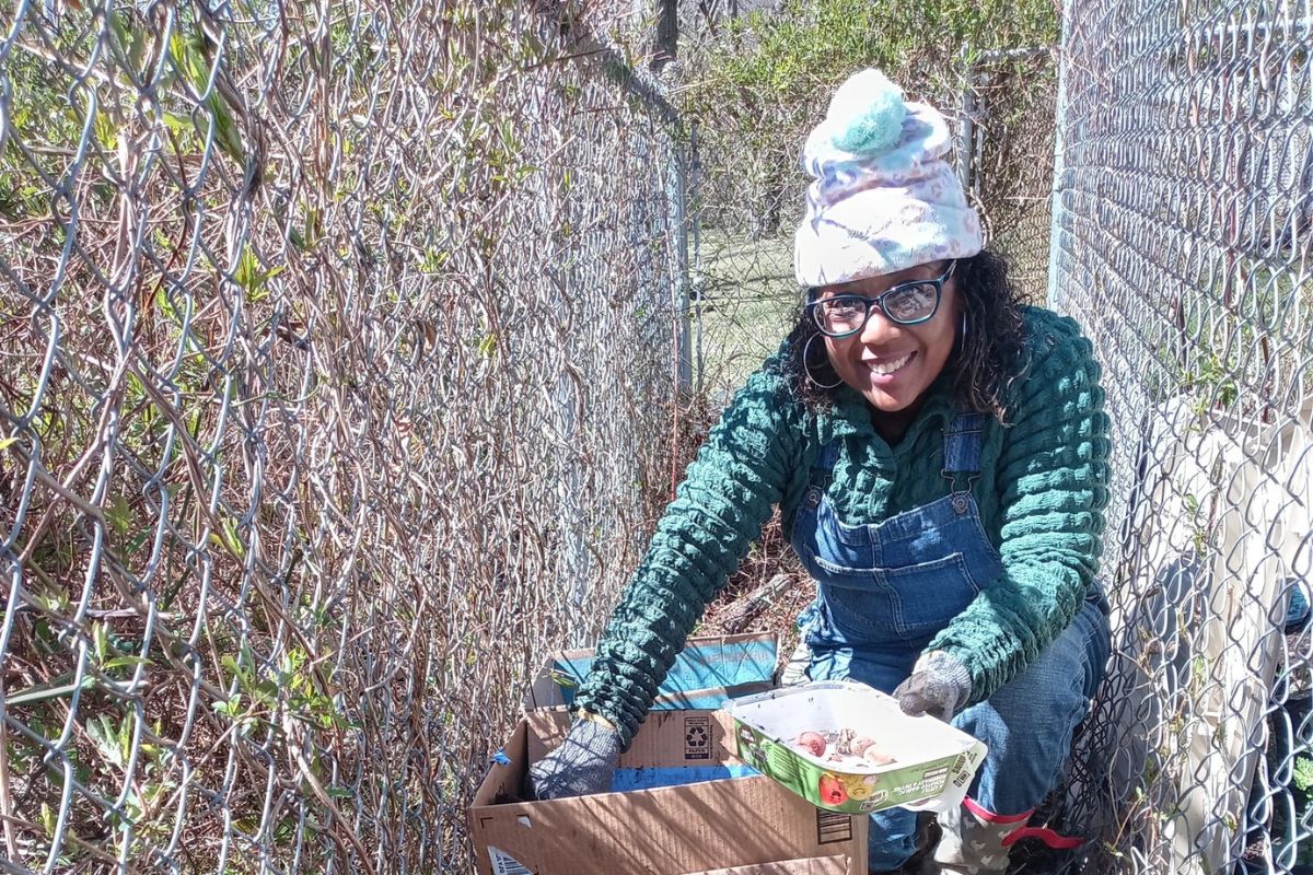 A person wearing glasses, a winter hat, and green sweater crouches by a chain-link fence, holding a tray of seeds or plants and packing items into a cardboard box.
