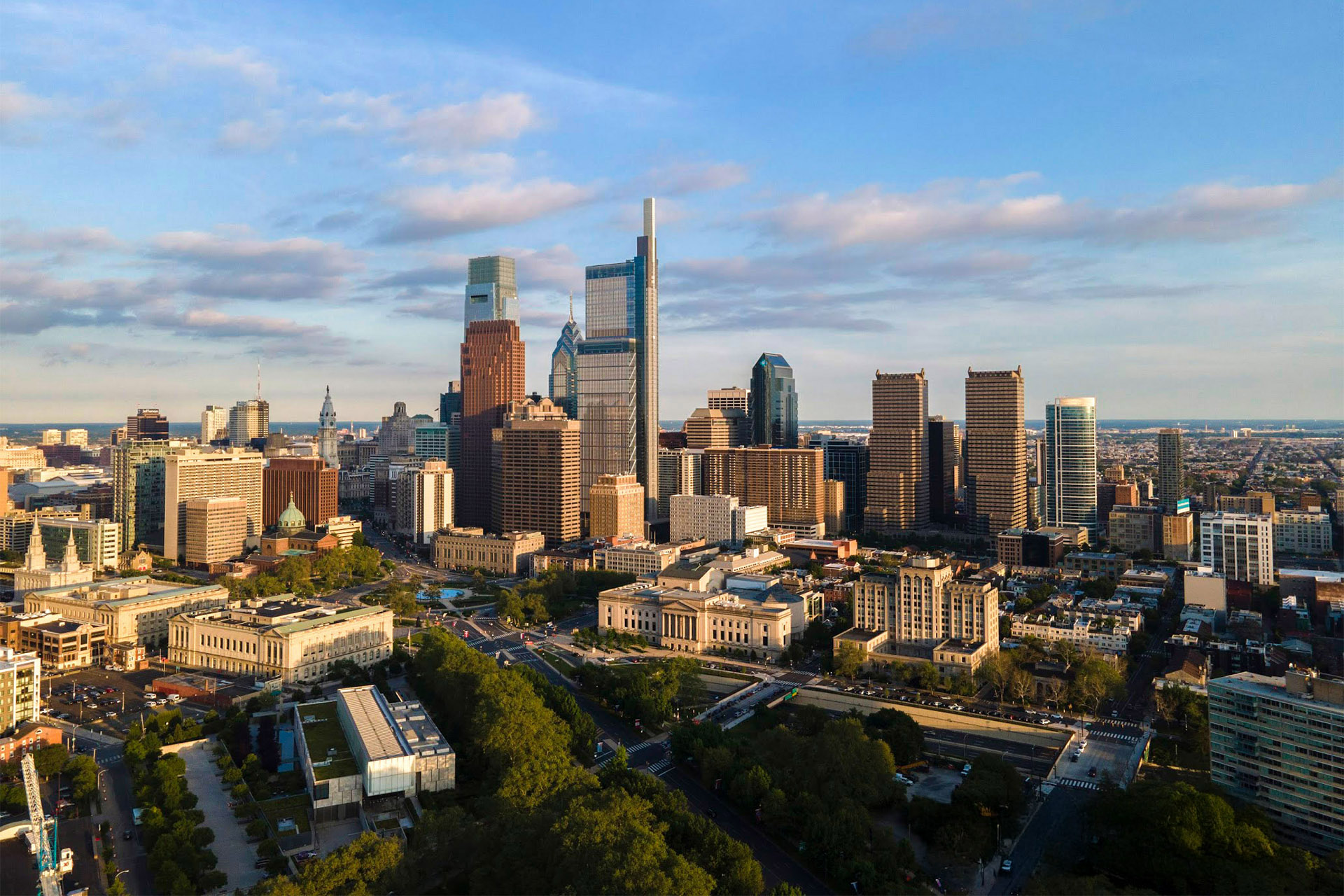 Aerial view of a downtown city skyline with tall modern buildings, tree-lined streets, and a mix of historic and contemporary architecture under a partly cloudy sky.