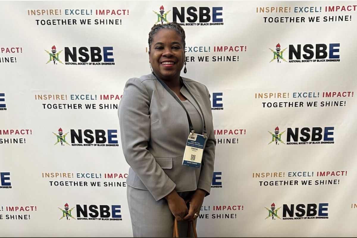 A person in a gray suit stands smiling in front of a backdrop with NSBE logos and the text "Inspire! Excel! Impact! Together We Shine!.