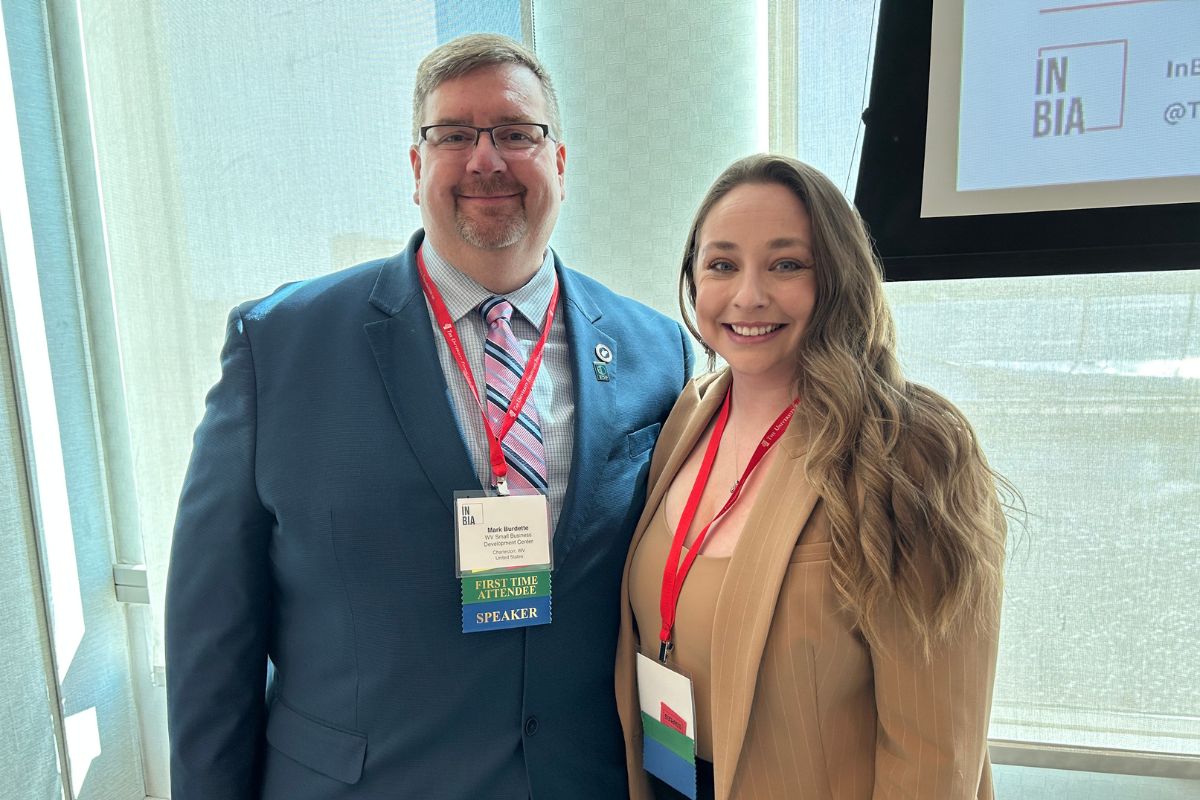 Two people smiling at a conference. The man wears a blue suit and a badge labeled "Speaker." The woman wears a tan jacket. Both have red lanyards. Large windows in the background.
