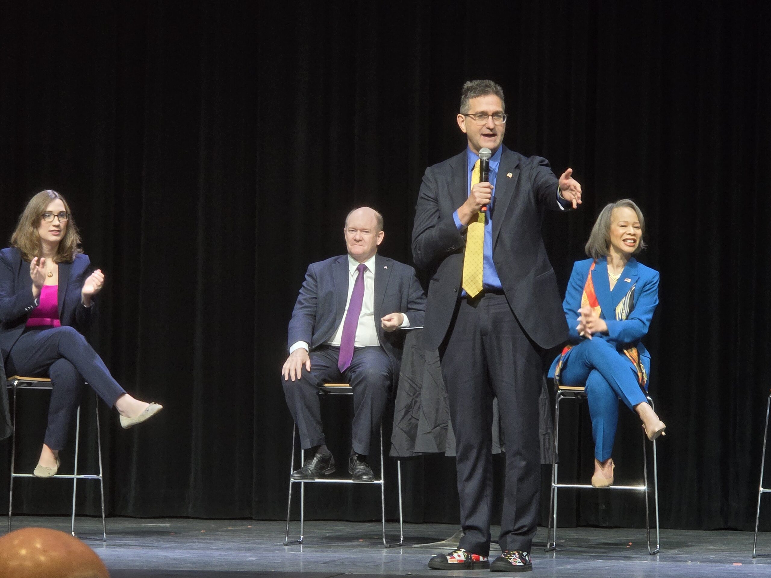 Four people in business attire are on a stage; one man stands speaking into a microphone while the others sit on stools against a black curtain backdrop.