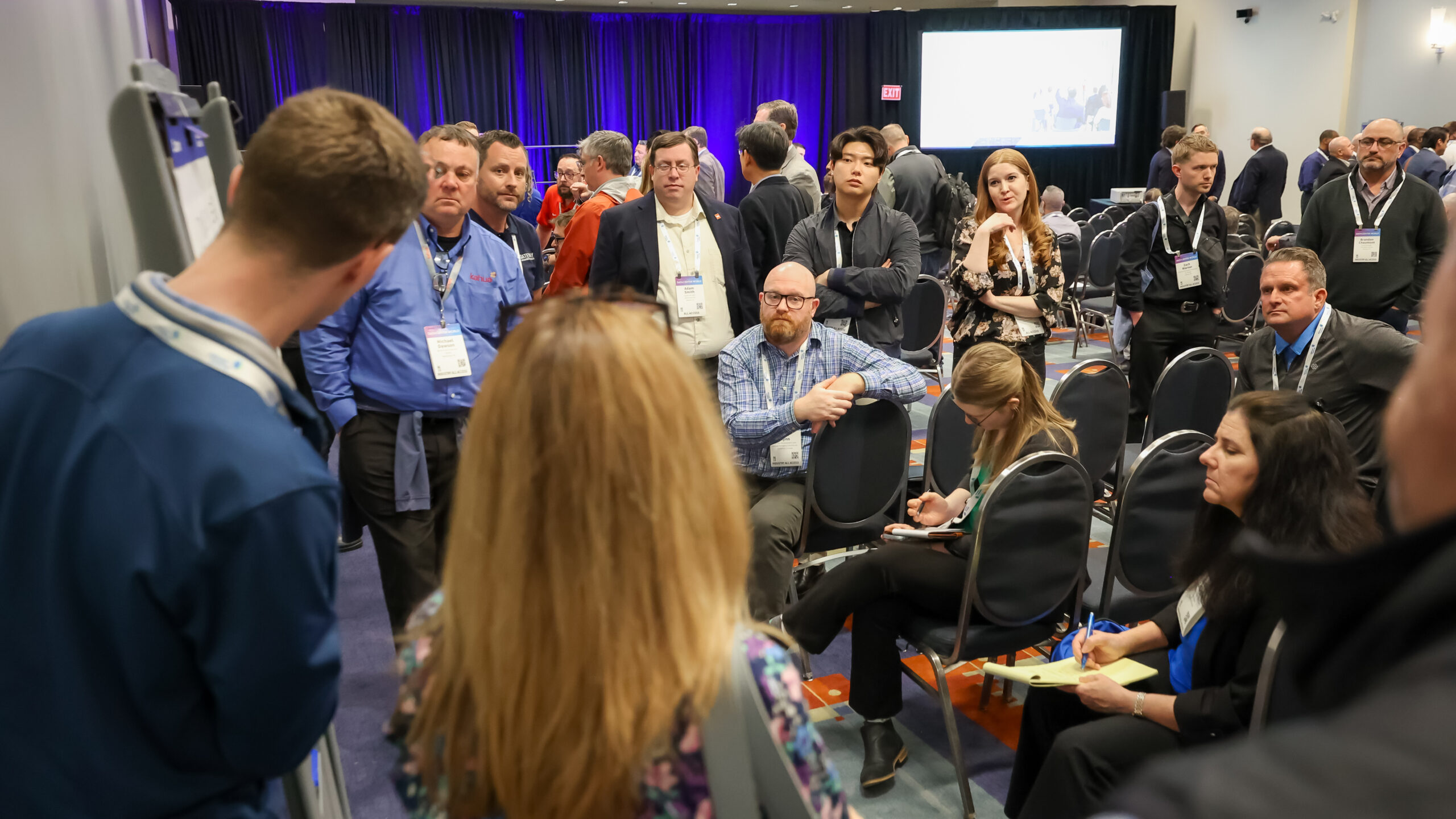 A group of people at a conference gather in a circle, some standing and some sitting, while listening and taking notes.