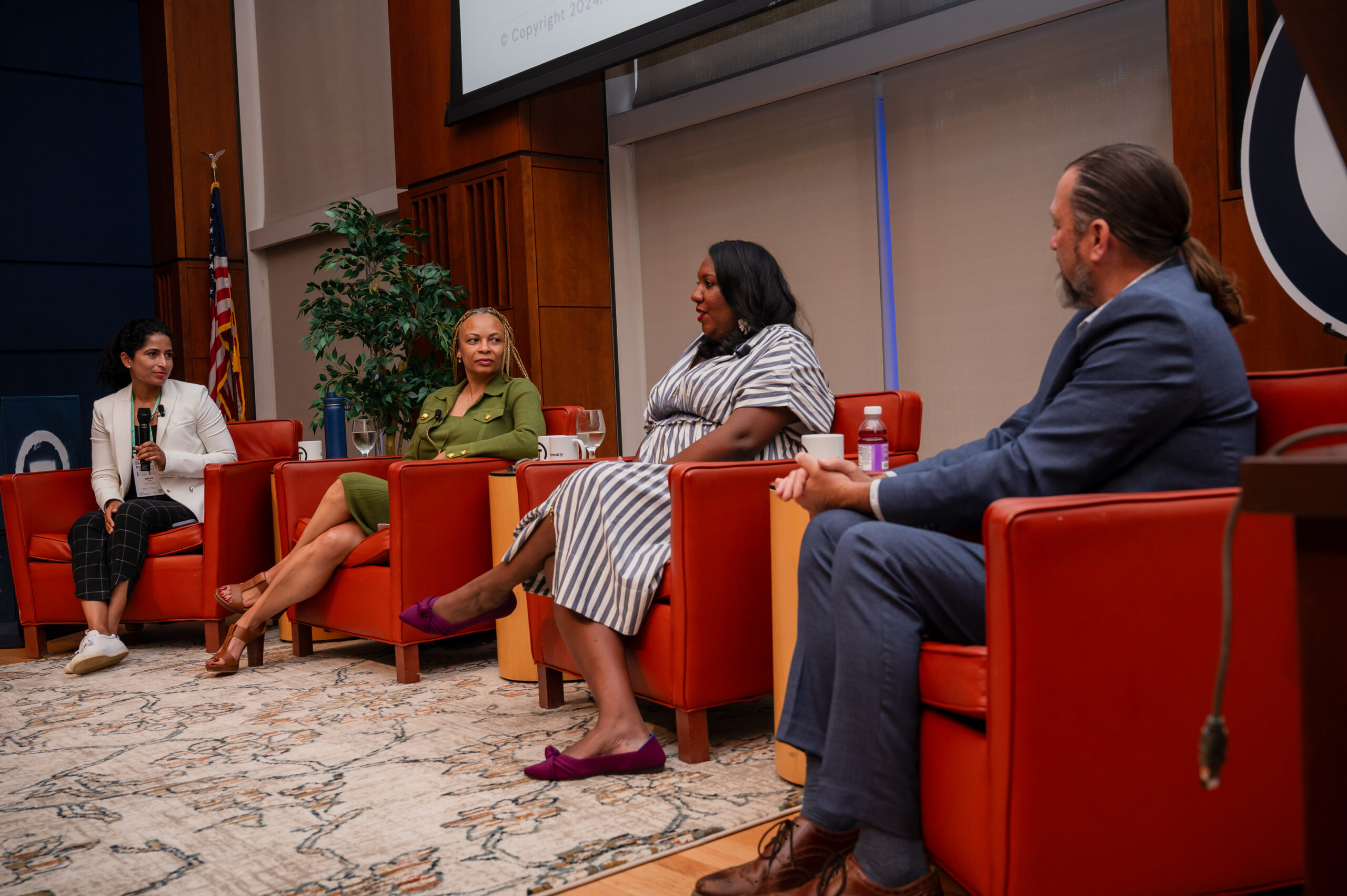 Four people sit on red chairs, engaged in a panel discussion in a conference room with a projector screen and US flag in the background.