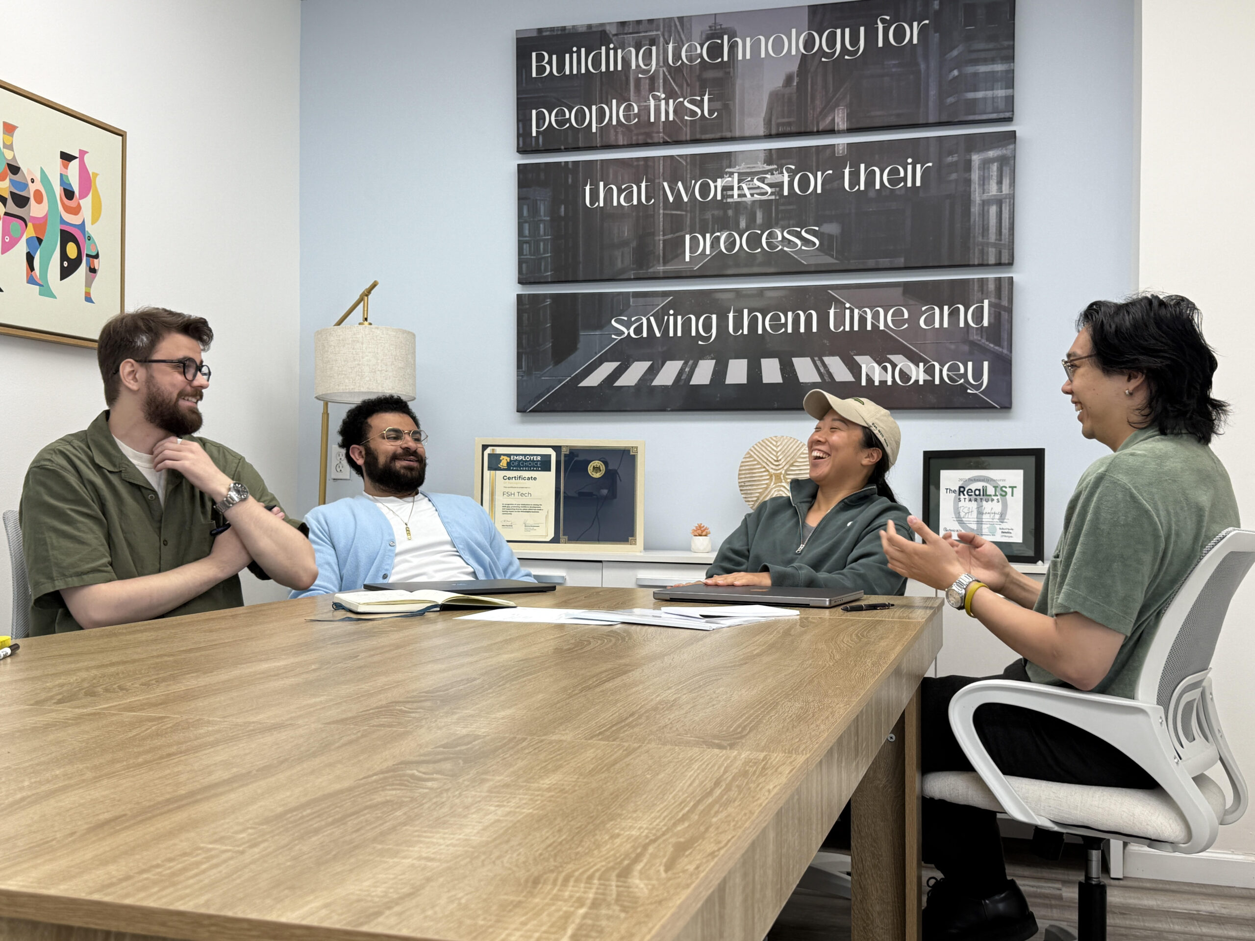Four people sit around a conference table engaged in discussion, with notepads, a laptop, and framed awards on the wall behind them.