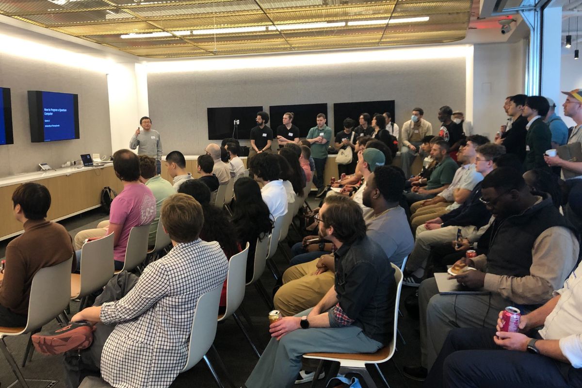 A group of people sit and stand in a modern conference room, listening to a speaker presenting slides on two large screens.