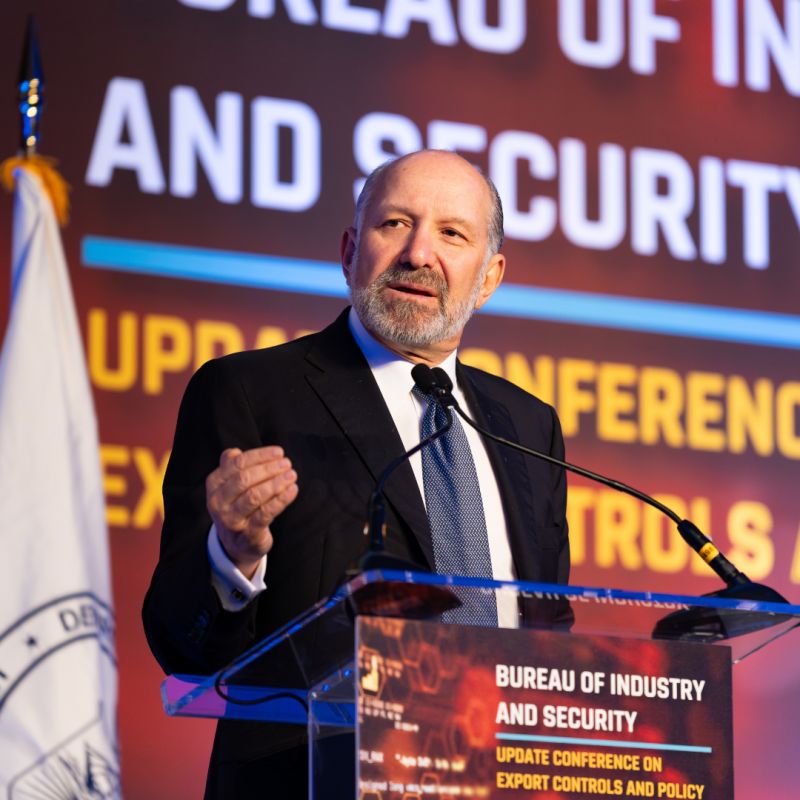 A man in a suit speaks at a podium labeled "Bureau of Industry and Security" during a conference on export controls and policy, with a government flag and screen in the background.