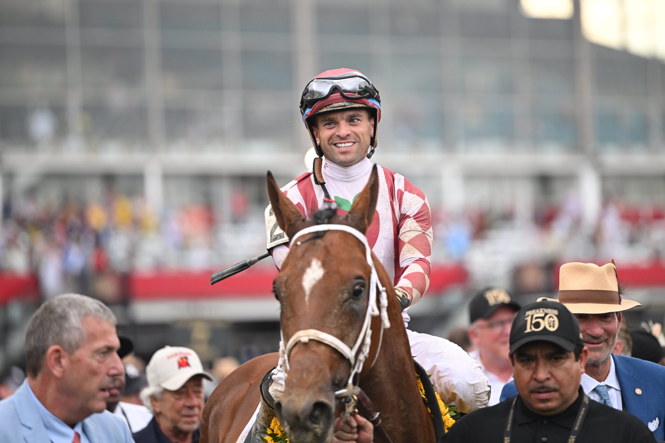 A jockey wearing a red and white uniform smiles while riding a brown horse in a crowded venue, surrounded by people.