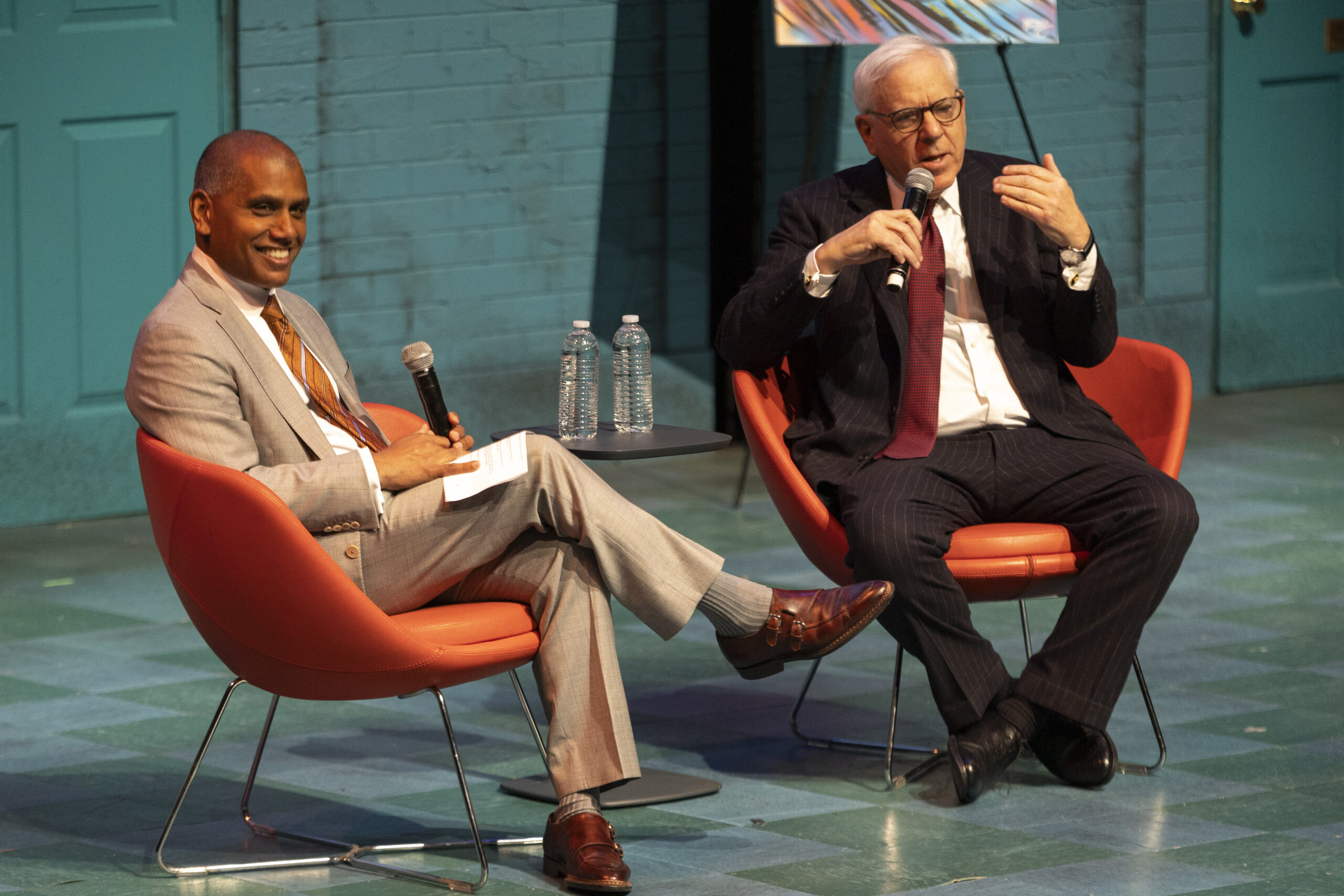 Two men in suits sit on stage in orange chairs, holding microphones and engaging in conversation; a small table with water bottles is between them.