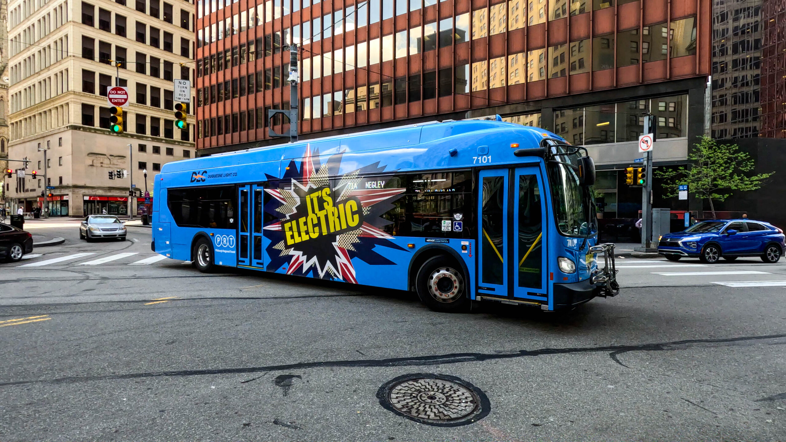 A blue city bus with "It's Electric" written on the side is turning at an intersection in an urban area with tall buildings in the background.