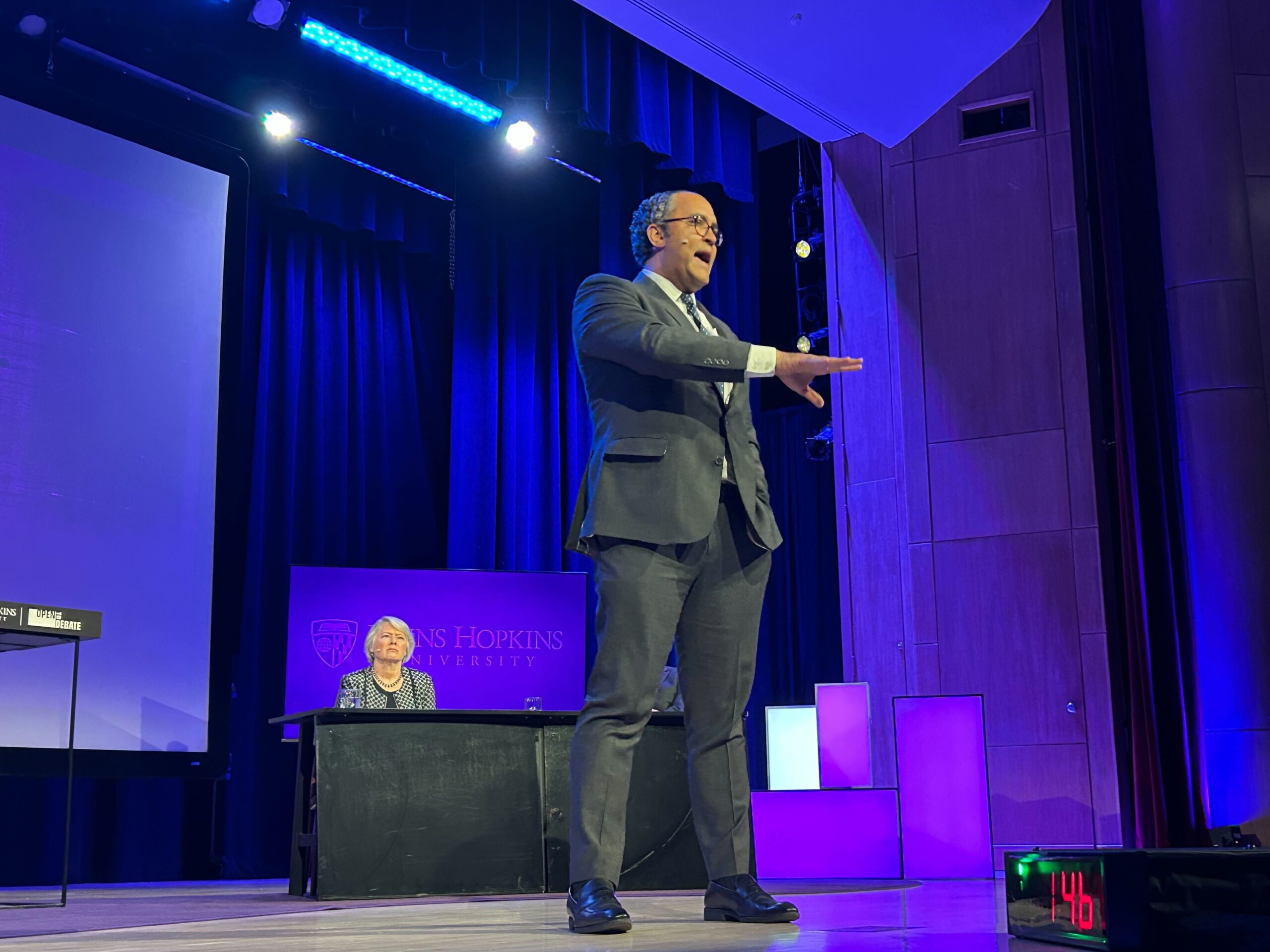 A man in a suit gestures while speaking on stage at Johns Hopkins University, with a seated person and a digital timer visible in the background.