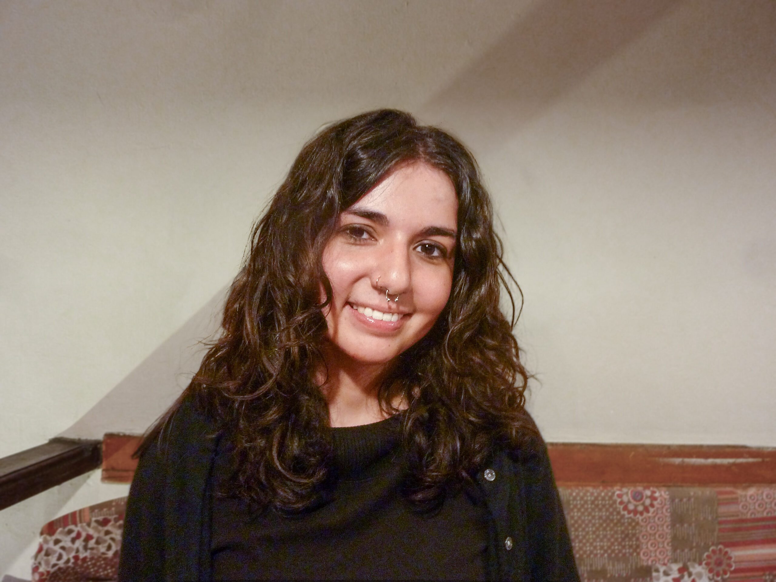 A young woman with long, wavy dark hair and a black top is sitting indoors, smiling at the camera. The background includes a light-colored wall and patterned cushions.