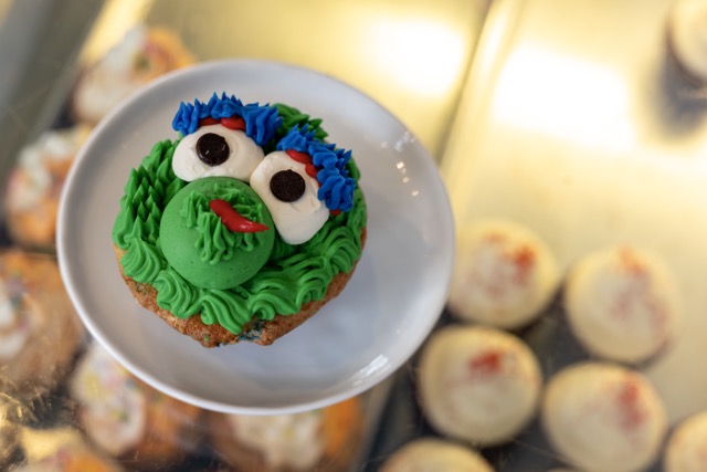 A cupcake decorated with green frosting, googly eyes, and a red mouth to resemble a cartoon face, placed on a white plate above other cupcakes.