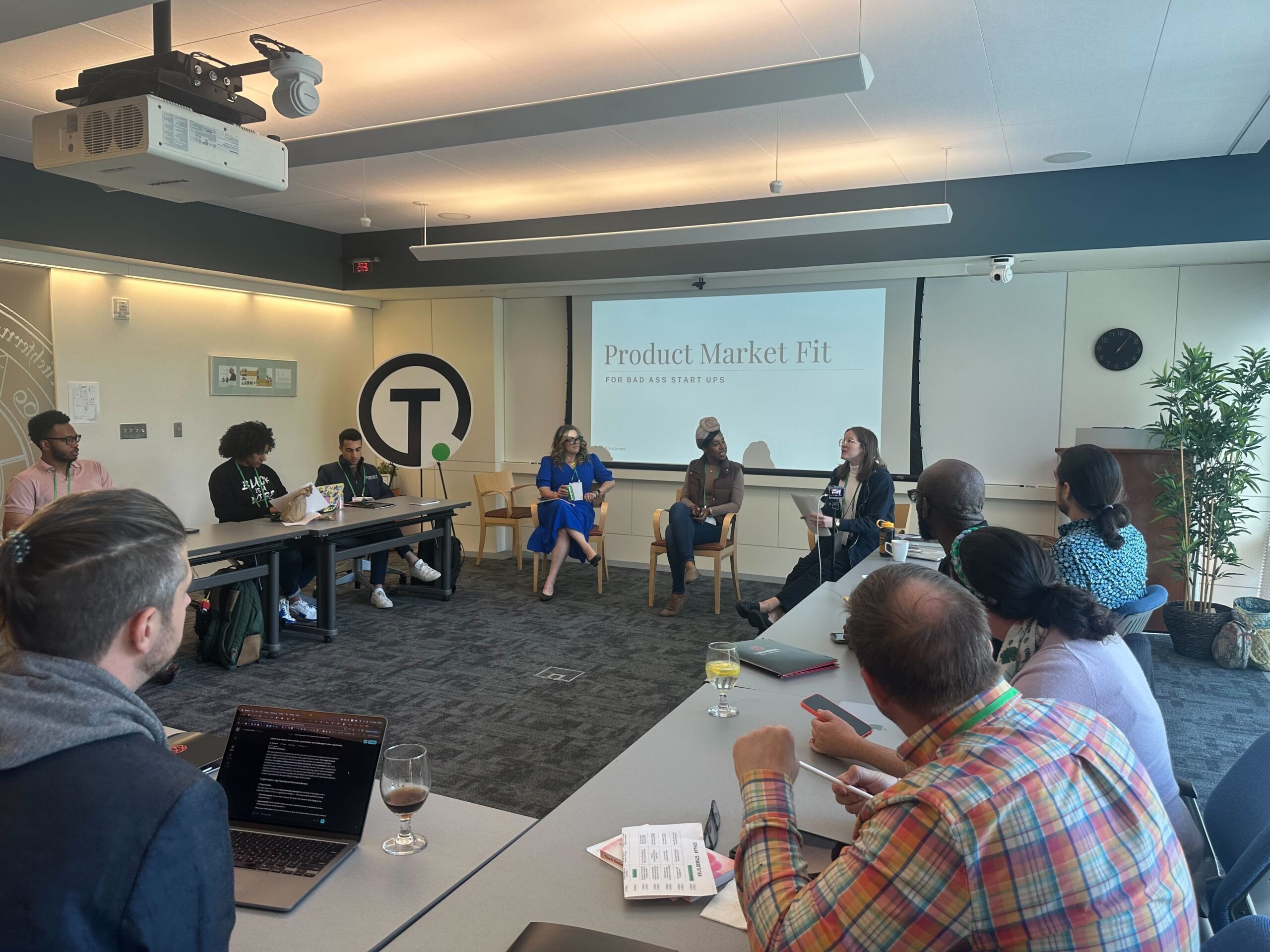 A panel of four people sits at the front of a conference room, leading a discussion on product market fit as others listen and take notes.