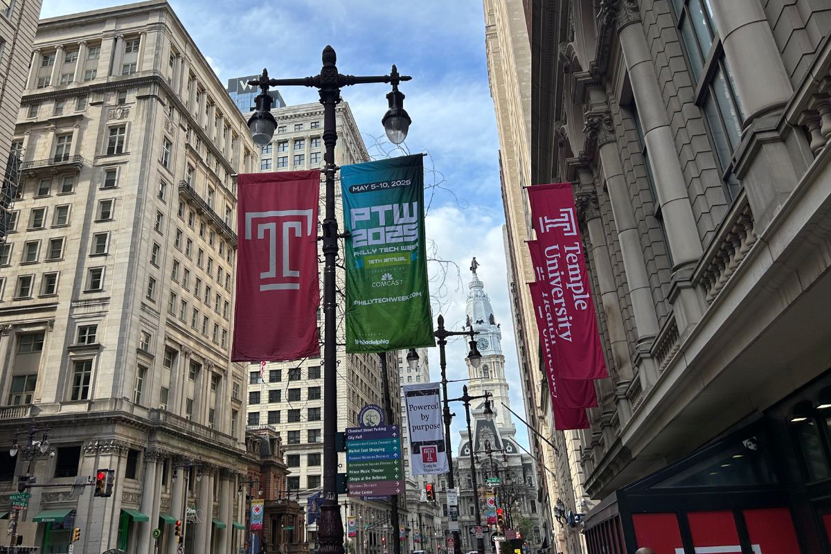 City street with banners for Temple University and PTW 2023 hanging from lamp posts, surrounded by tall buildings and the Philadelphia City Hall visible in the background.