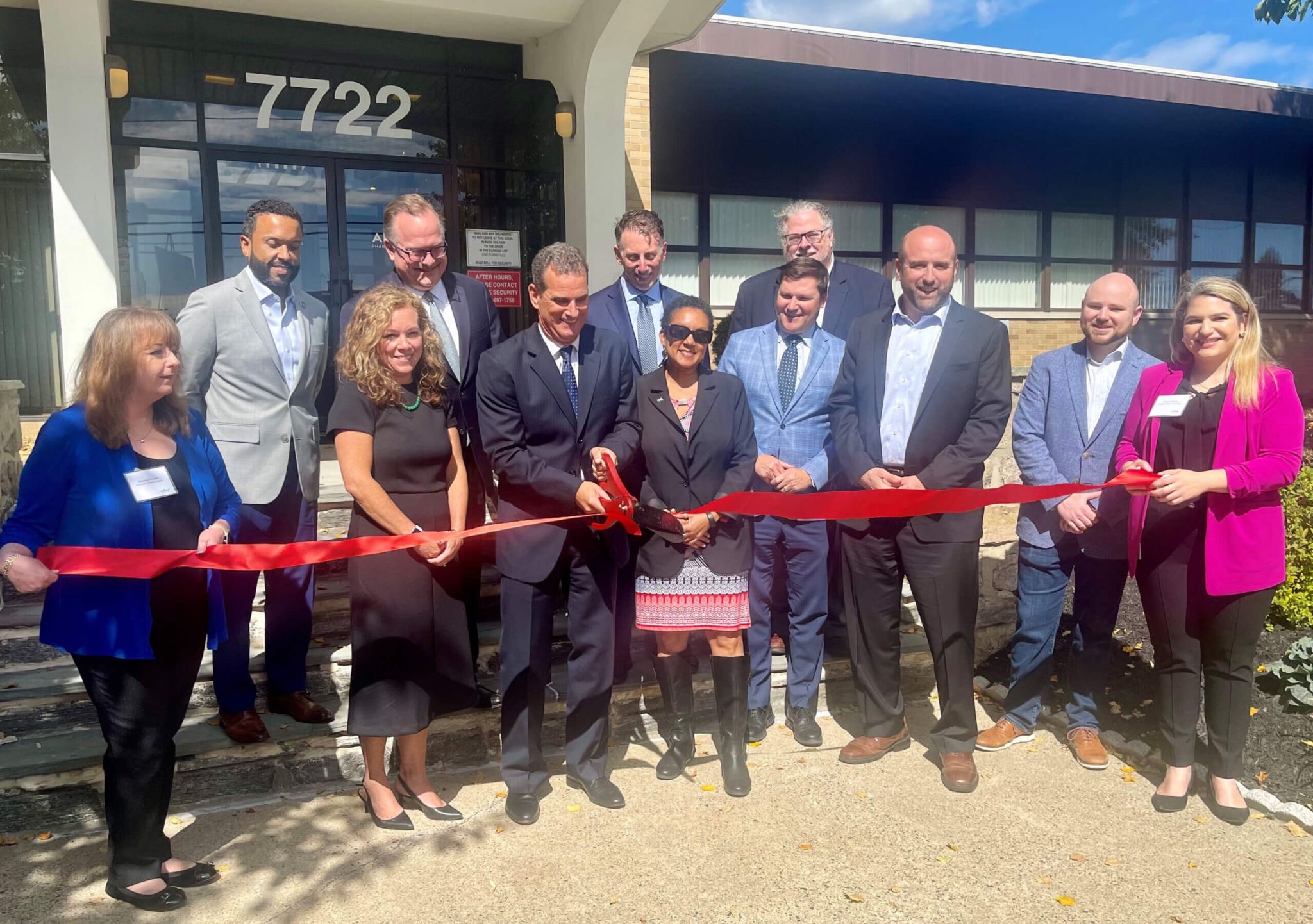 A group of people in business attire stand outside a building, holding and cutting a red ribbon for a ribbon-cutting ceremony.