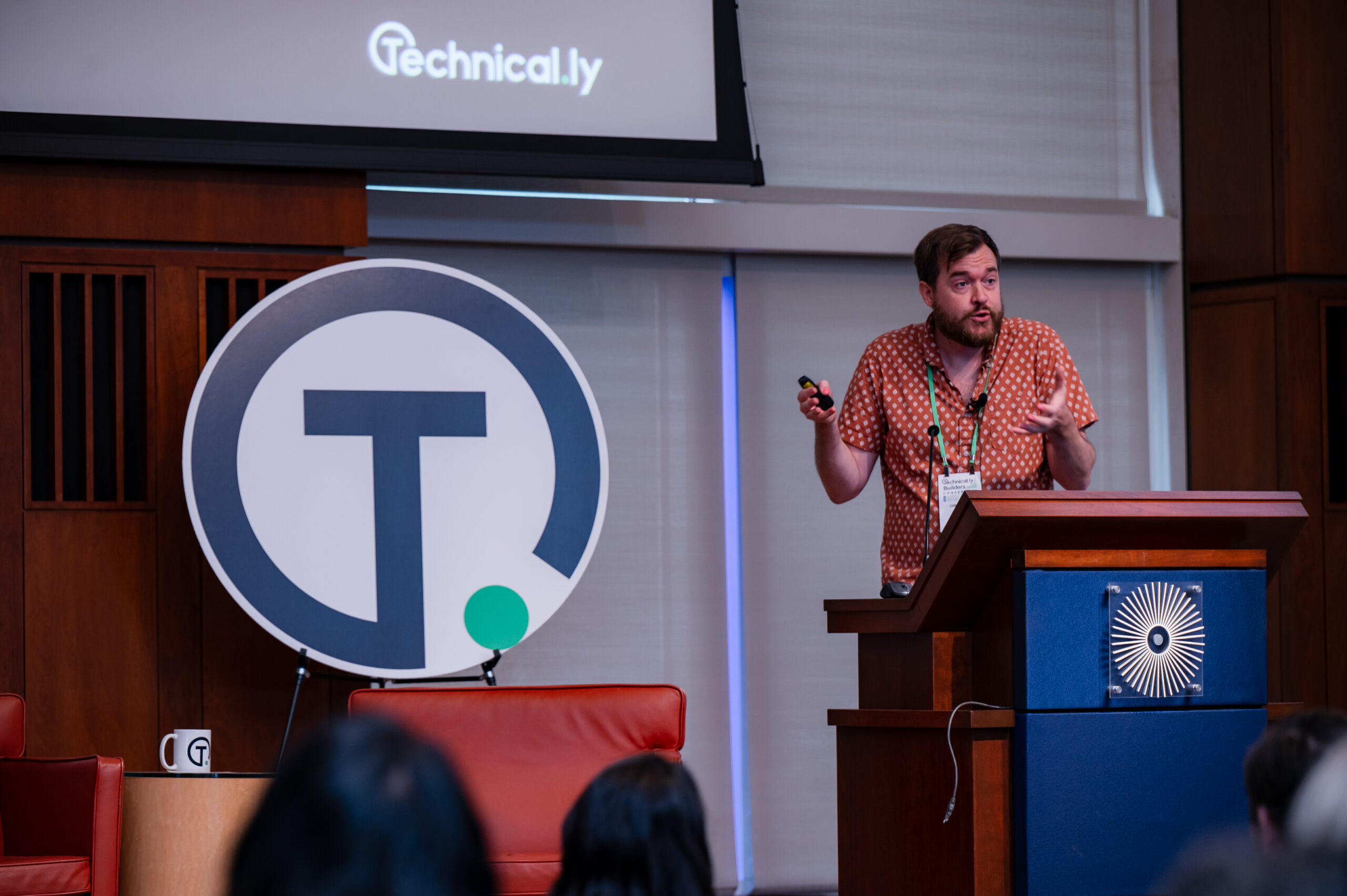 A man stands at a podium giving a presentation, with a large sign displaying a "T" and a projection screen showing the Technical.ly logo in the background.