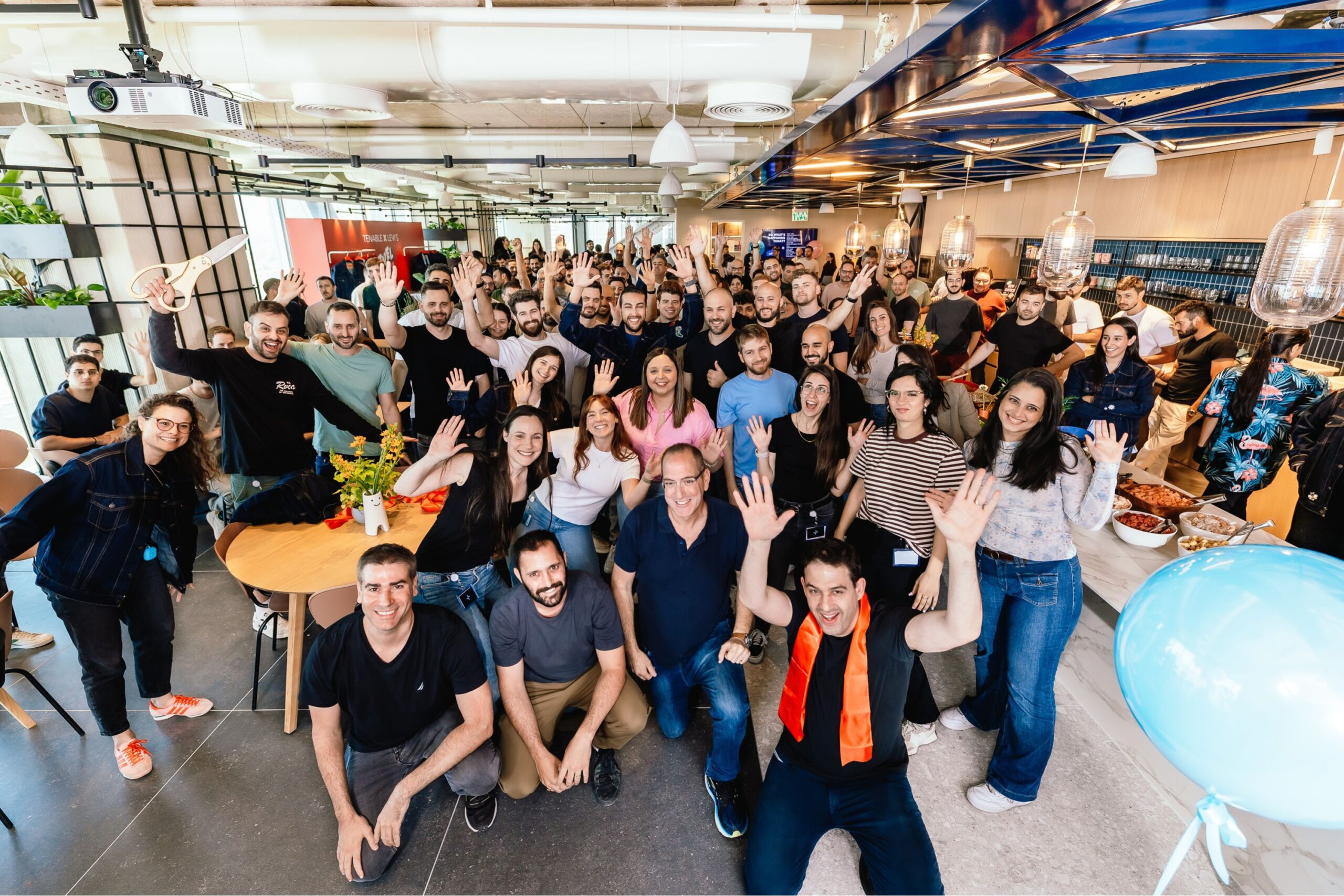 A large group of people gathered indoors, many smiling and waving at the camera in a modern, brightly lit office space.