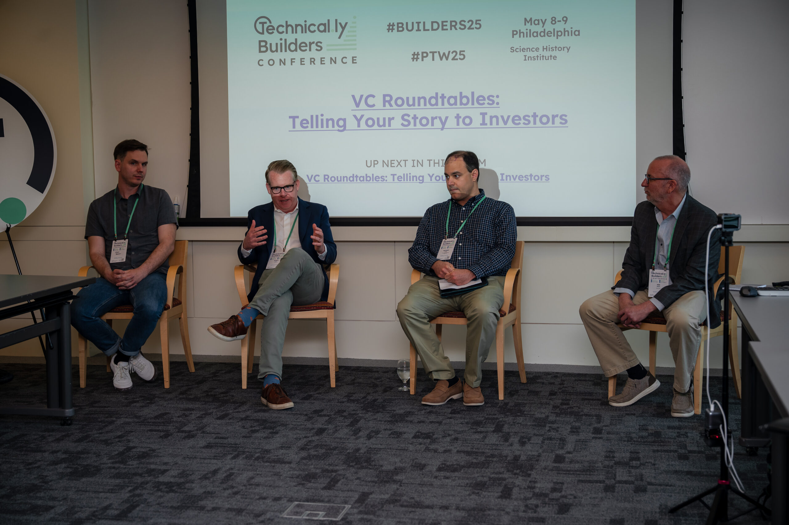Four men sit on a panel discussion at a conference, with a screen behind them displaying information about the event and upcoming session.