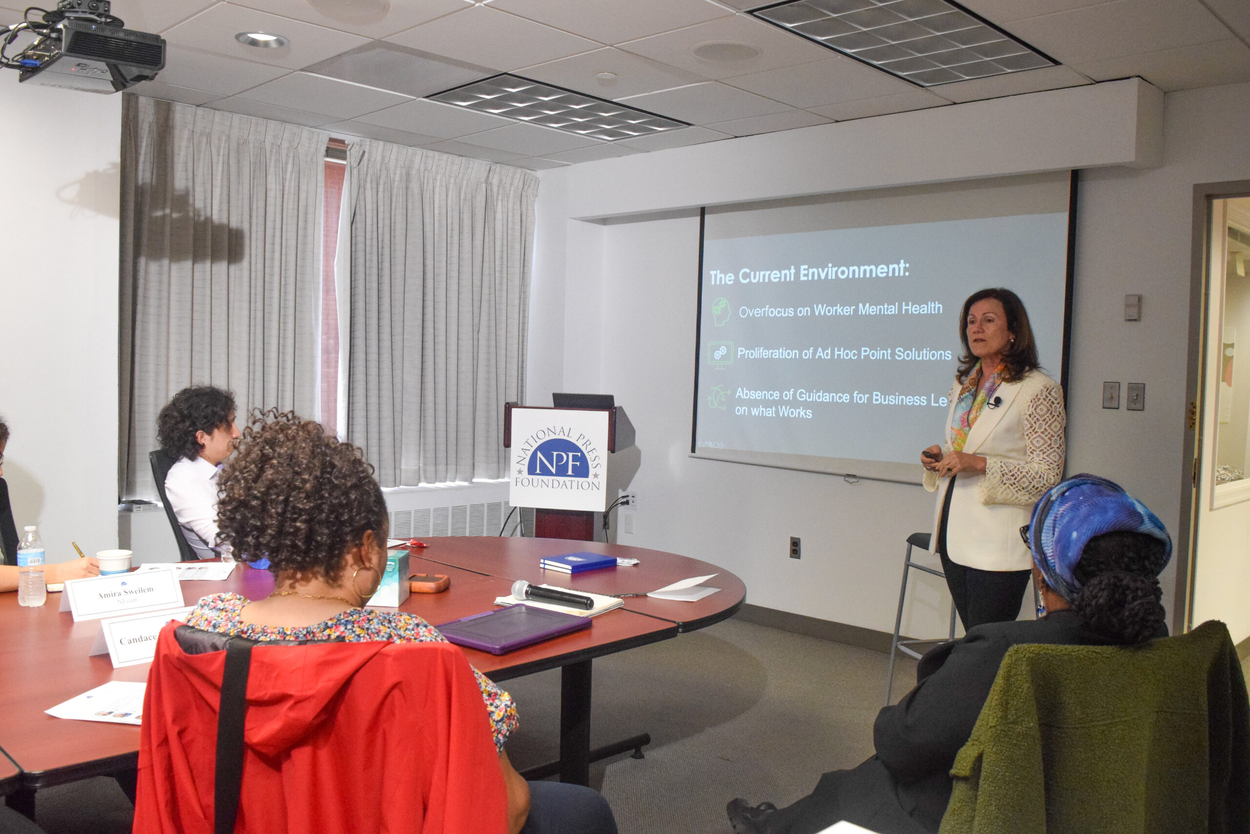 A woman stands and presents a slideshow to a small seated group in a conference room; the slide discusses trends in workplace mental health.
