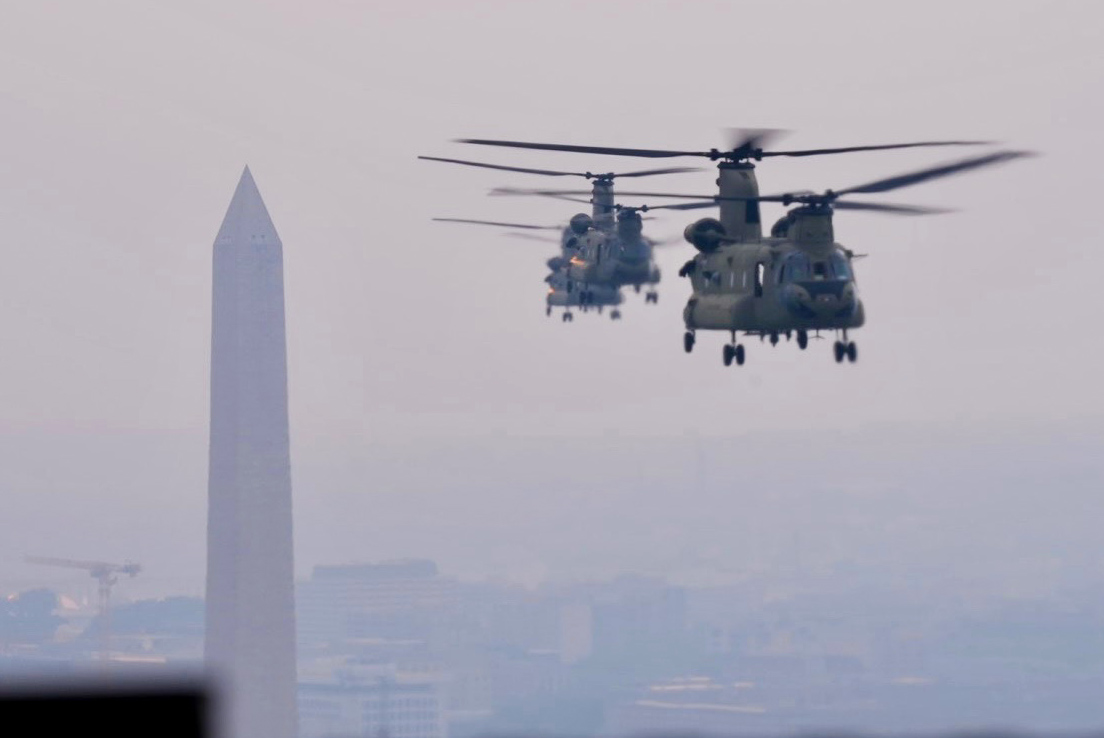 Three military helicopters fly near the Washington Monument in hazy weather, with city buildings faintly visible below and in the background.
