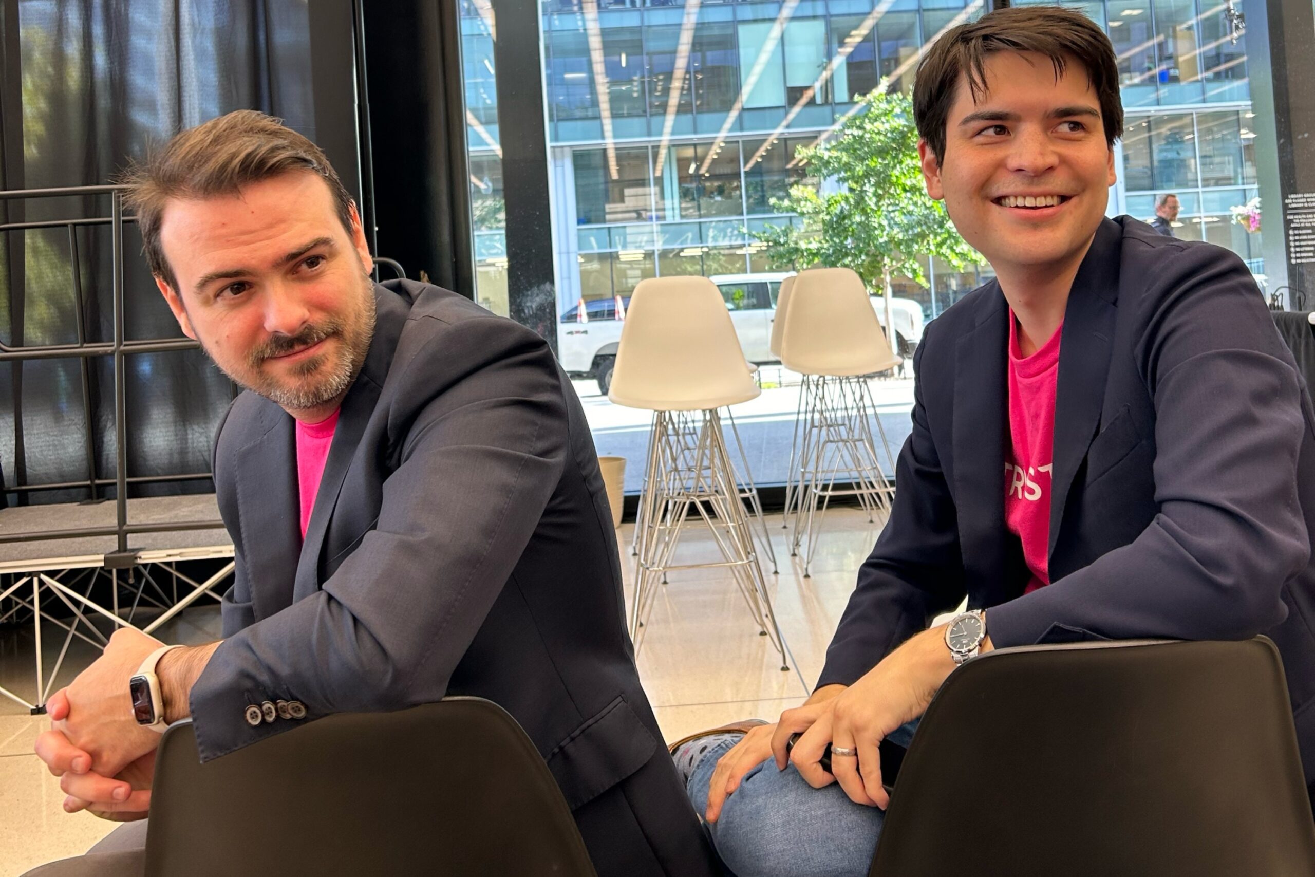 Two men in blazers and pink shirts sit back to back on black chairs indoors, with empty white chairs and large windows in the background.