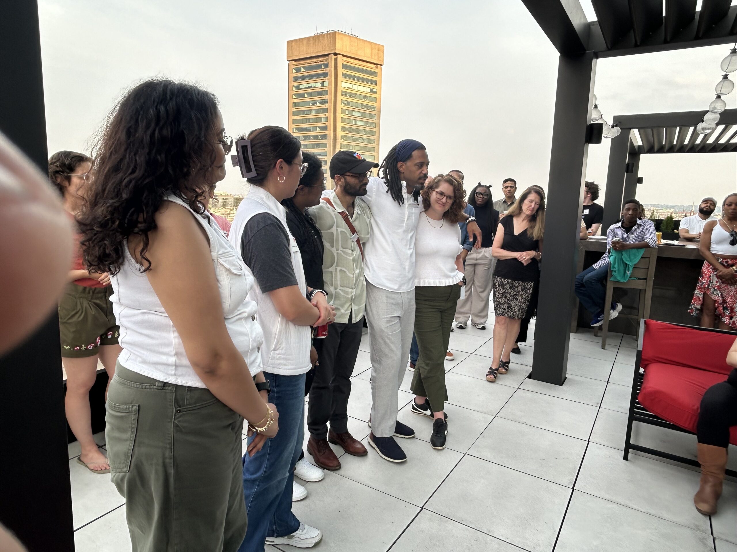 A group of people gathered on a rooftop patio, some standing and some seated, with a tall building in the background.