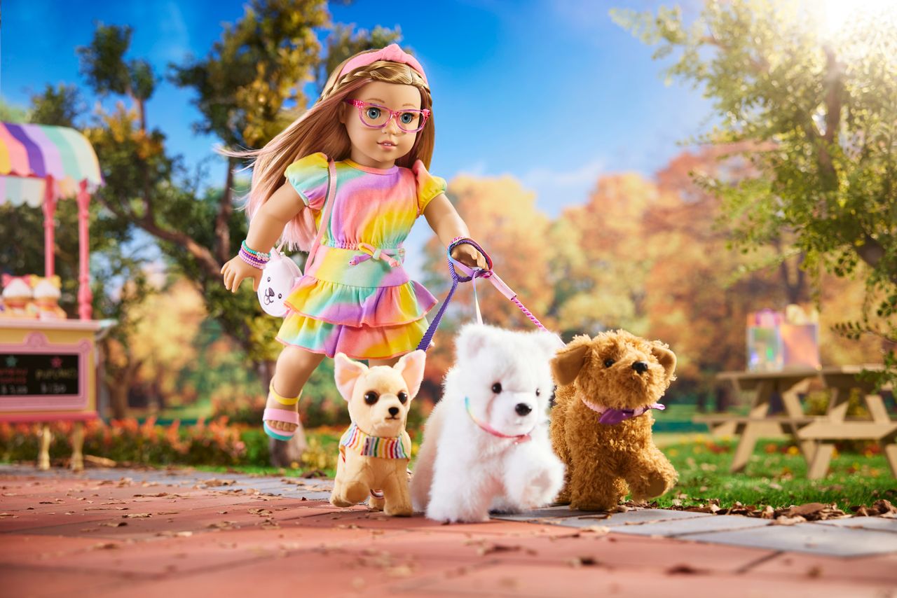 A doll in a colorful dress and glasses walks three plush dogs on leashes in a park setting with autumn trees and a picnic table in the background.