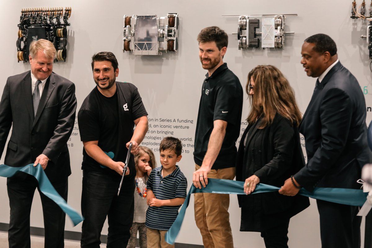 Six adults and two young children participate in a ribbon-cutting ceremony in front of a wall display featuring mechanical devices.