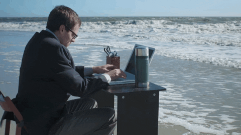 A man in a suit works on a laptop at a desk set up on the shoreline, with waves approaching in the background.