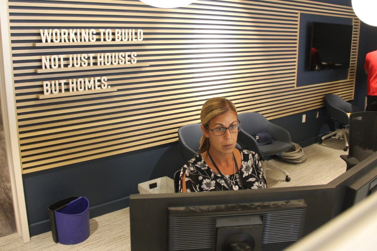 A woman works at a computer in a modern office with a sign on the wall that reads, "WORKING TO BUILD NOT JUST HOUSES BUT HOMES.