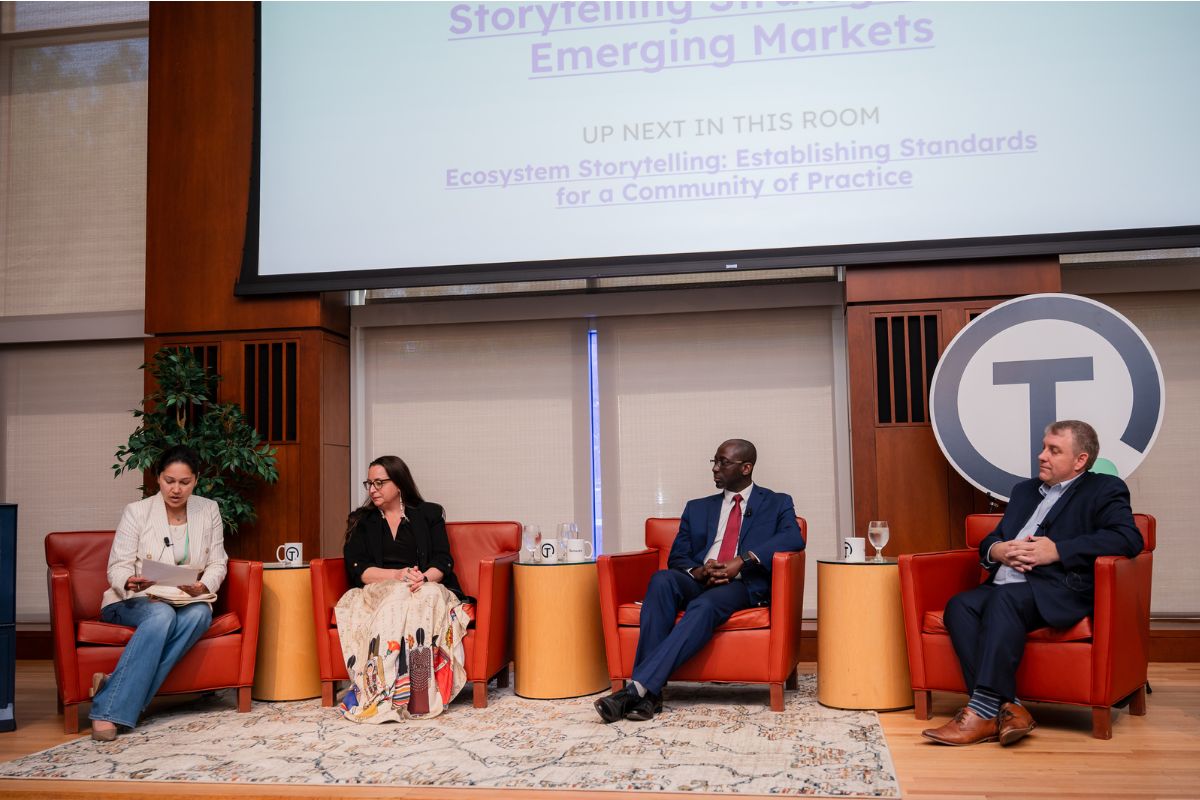 Four panelists sit on a stage in orange chairs, engaged in discussion, with a screen behind them displaying the session title and agenda.