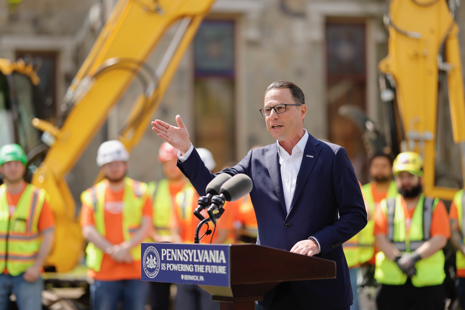 A man speaks at a podium labeled "Pennsylvania is powering the future," with construction workers and equipment visible in the background.