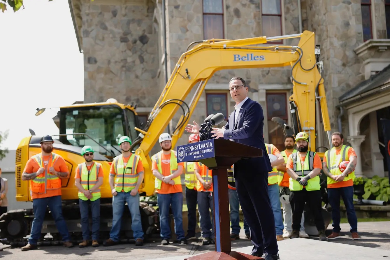 A man speaks at a podium labeled "Pennsylvania" in front of construction workers and a yellow excavator at a site with a stone building in the background.