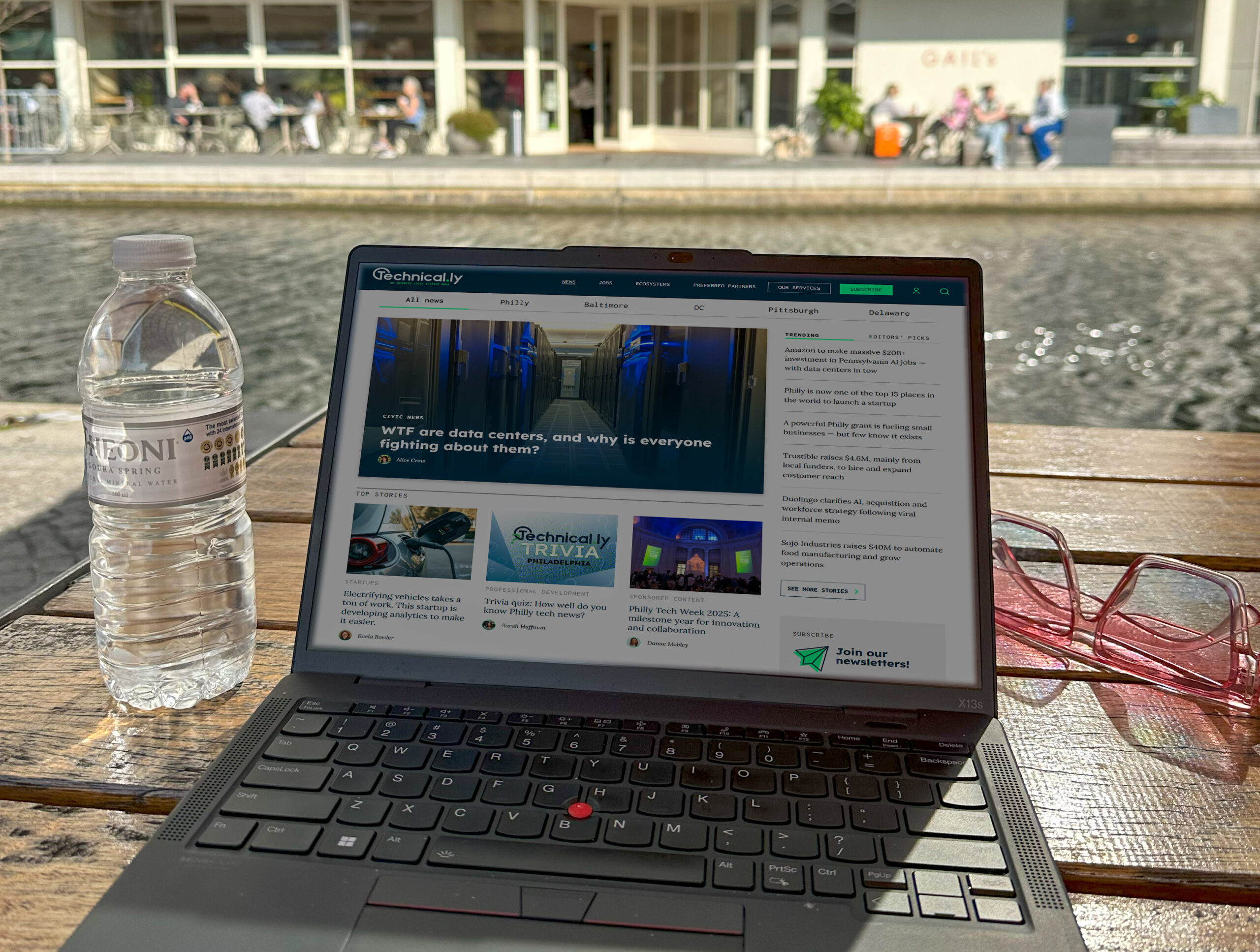 A laptop on a wooden table displays a tech news website next to a bottle of water and red glasses, with a pond and modern building in the background.