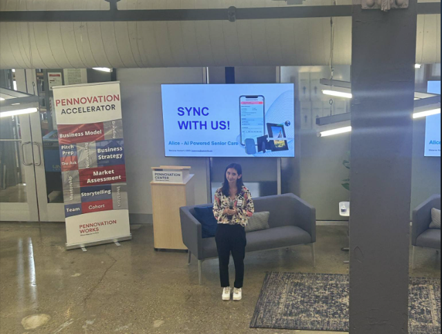 A woman stands and speaks in front of a screen displaying “SYNC WITH US! Ailce: AI Powered Senior Care,” in a modern office with Pennovation Accelerator signs.
