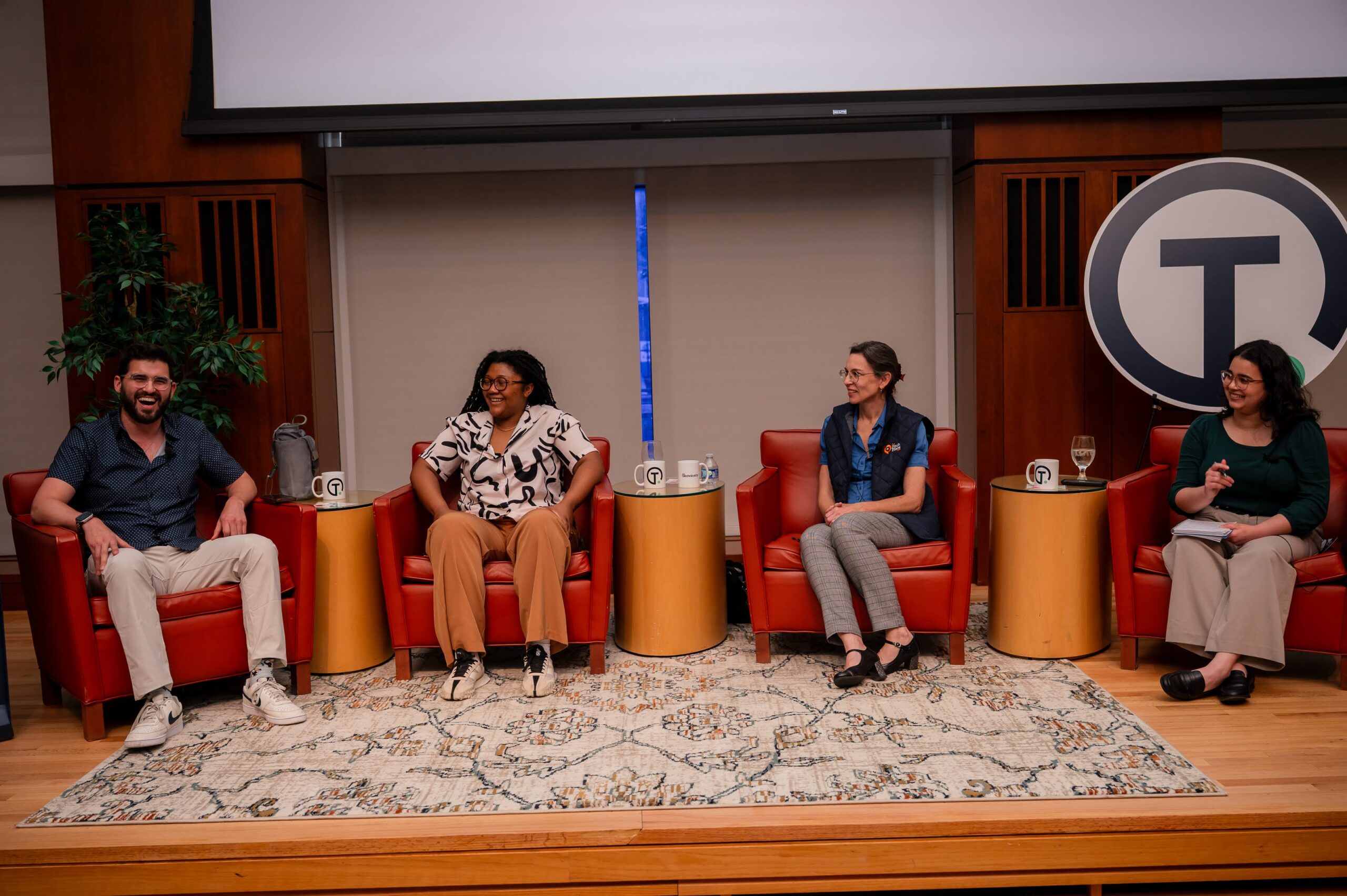 Four people sit in red chairs on a panel discussion stage, with a rug underfoot, mugs and water on tables, and a large circular logo in the background.
