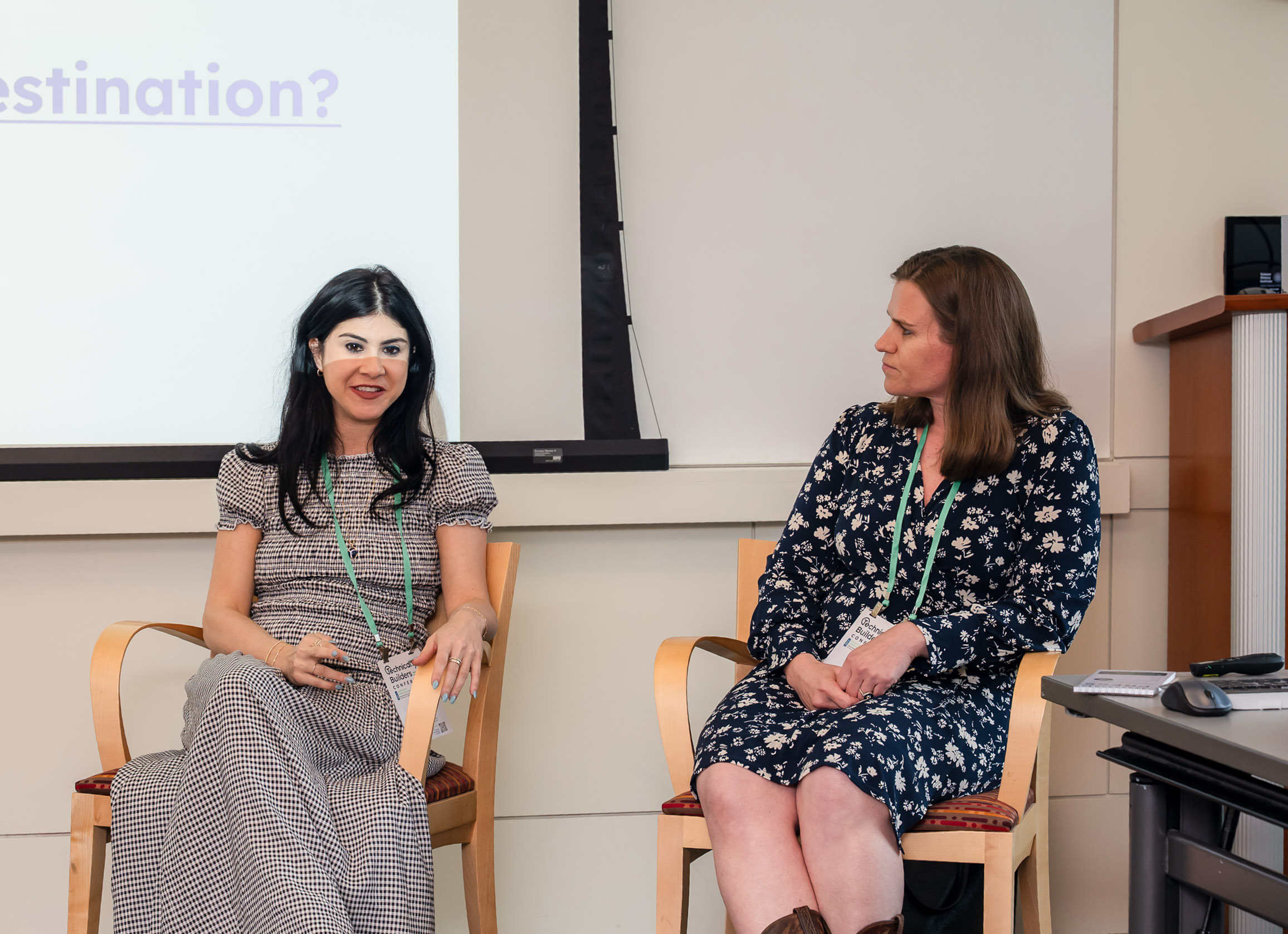 Two women sit and talk at a panel discussion; one is speaking while the other listens. A projection screen and conference lanyards are visible.