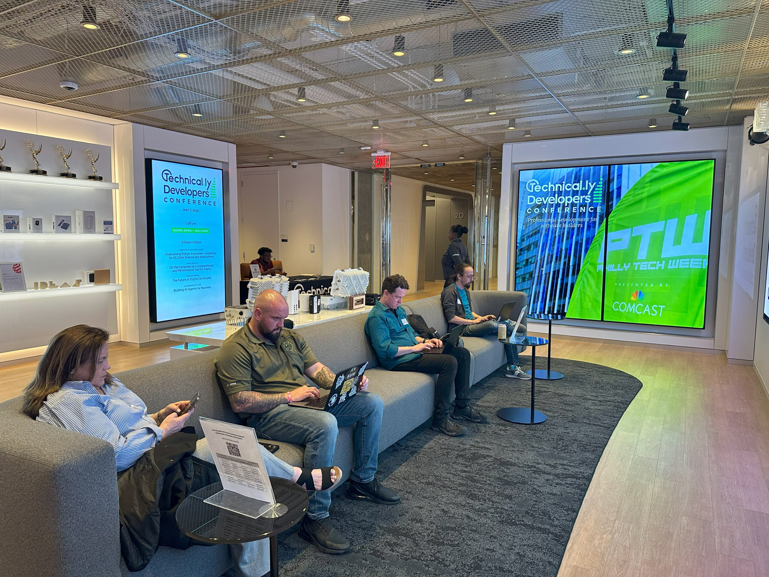 Four people sit on couches using laptops and phones in a modern lounge area at a tech conference, with large digital displays and awards visible in the background.