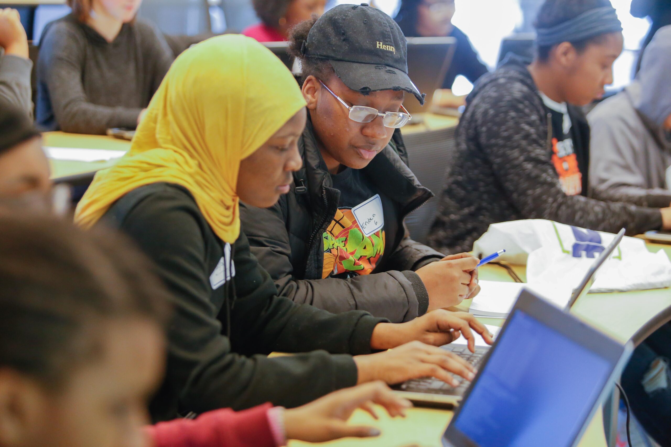 Two students sit at a desk working on laptops, with others visible in the background, in a classroom or workshop setting.