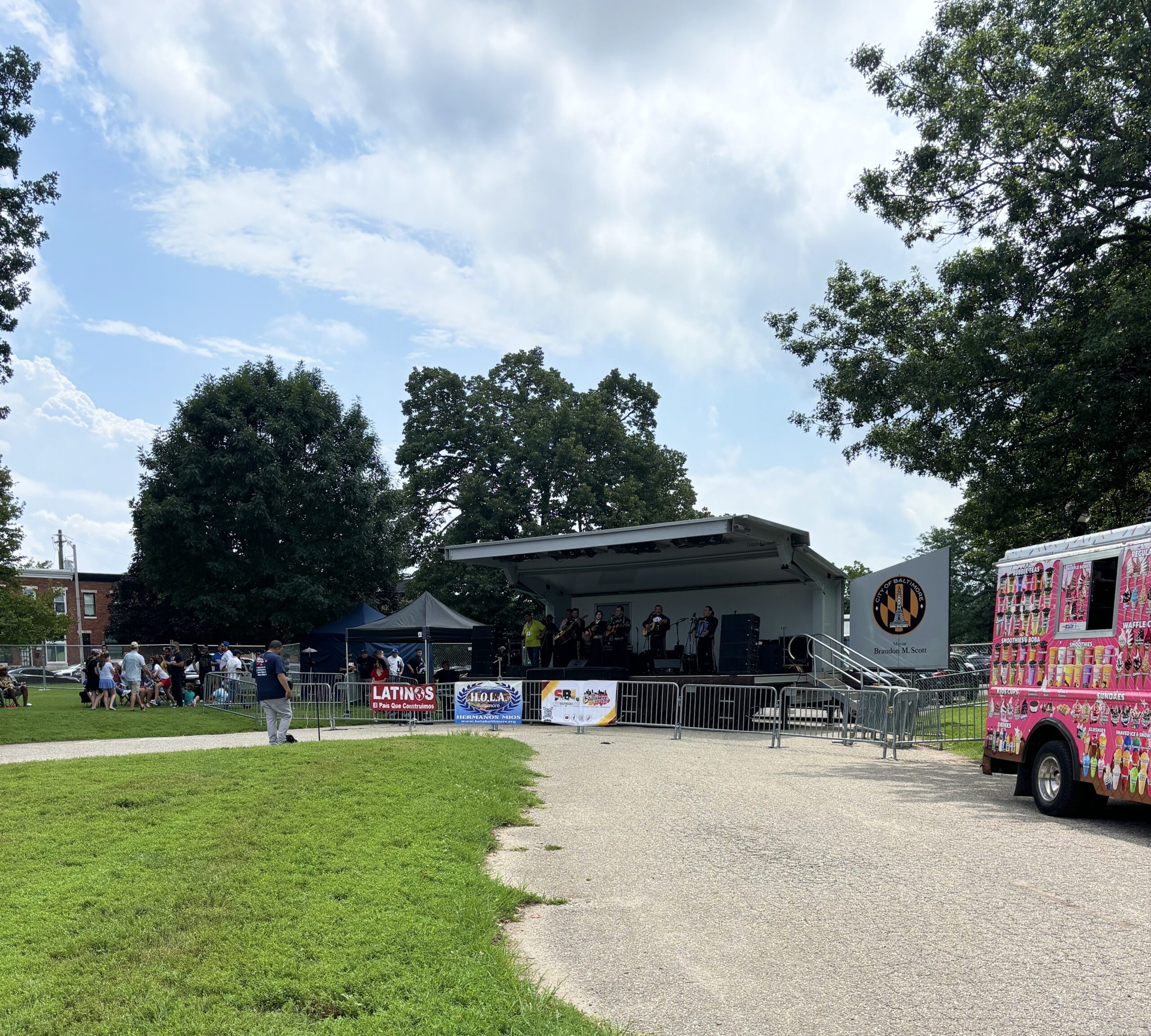 A band performs on an outdoor stage in a park with people sitting nearby. An ice cream truck is parked to the right, and event banners are displayed on the stage barriers.