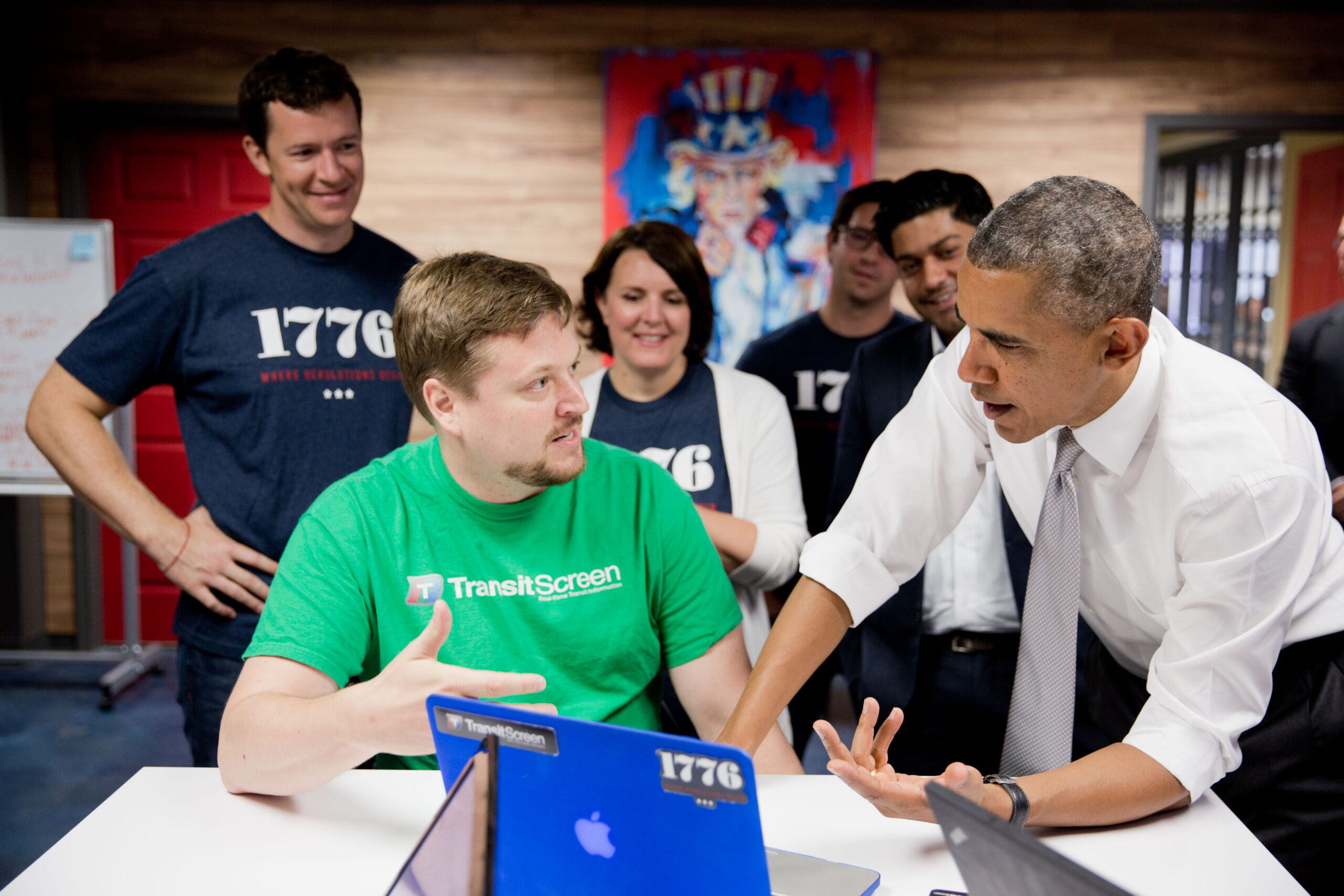 A group of people gathers around a table with laptops as a man in a white shirt and tie speaks to a seated man in a green shirt; others watch in the background.